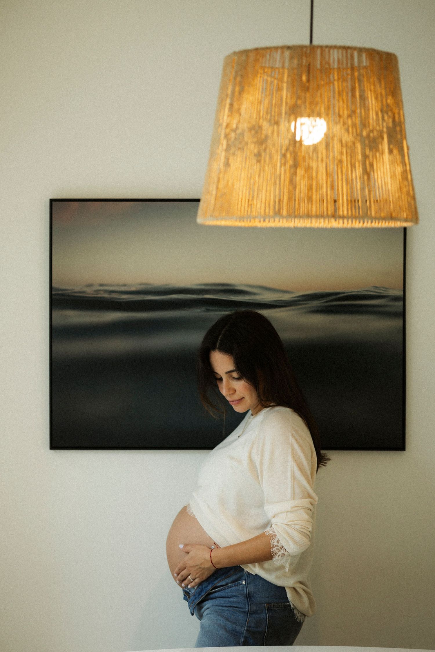 Pregnant woman in white top and jeans cradling baby bump, standing by ocean artwork under warm pendant light.