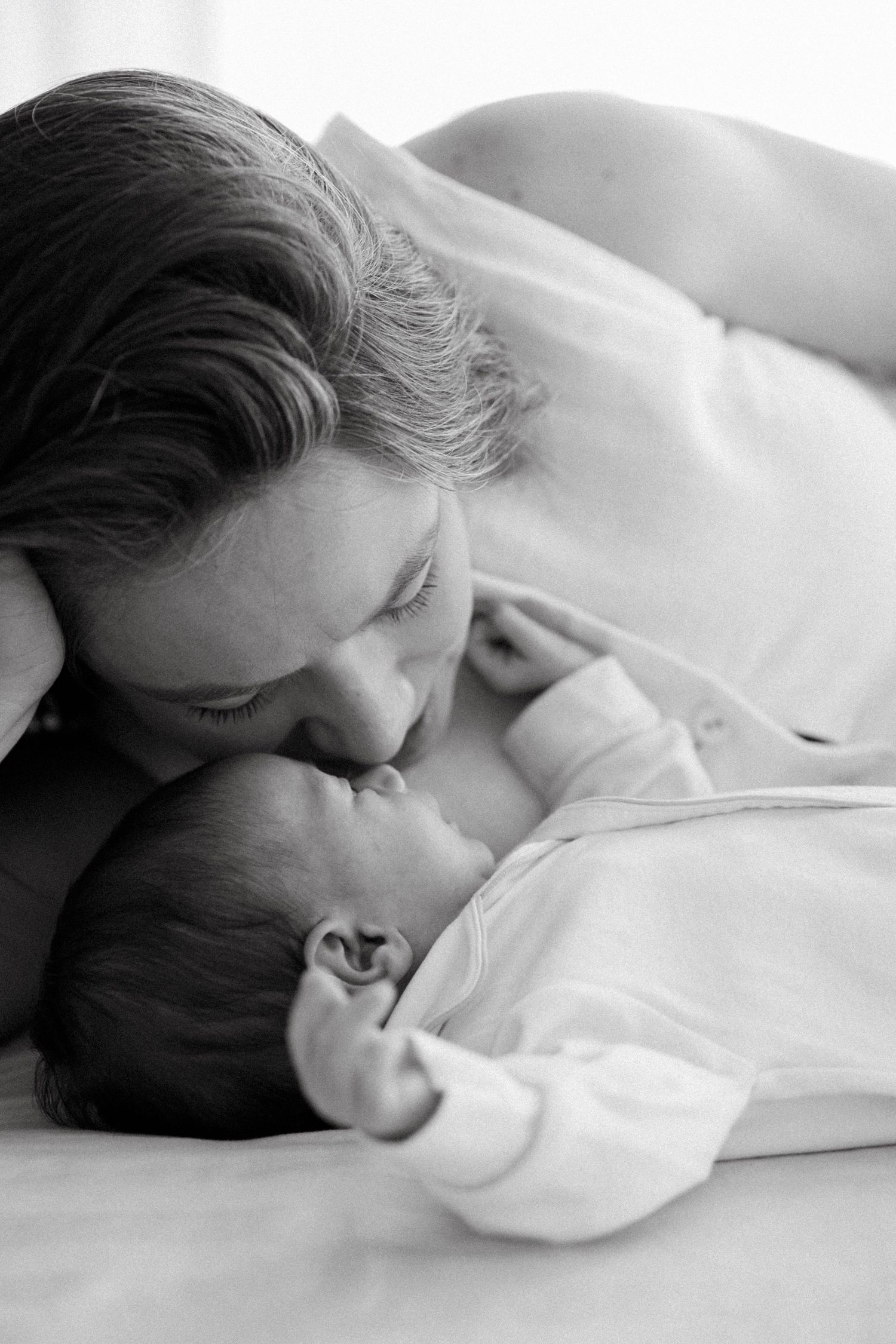 Black and white photo of a mother tenderly kissing her newborn baby while lying together on a bed.