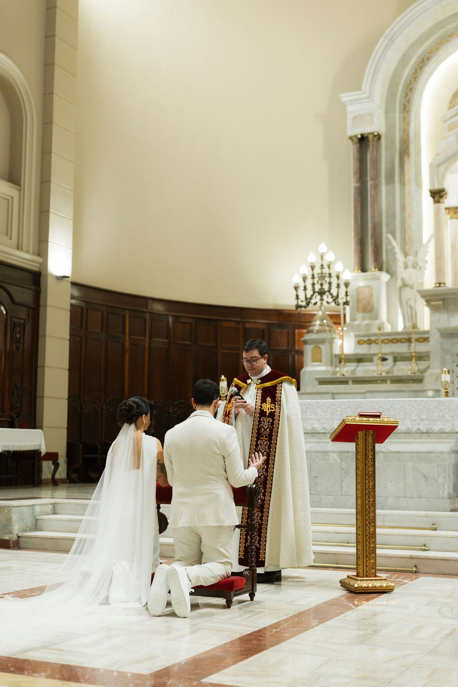Bride and groom kneeling before a priest at a church altar during a traditional Catholic wedding ceremony.