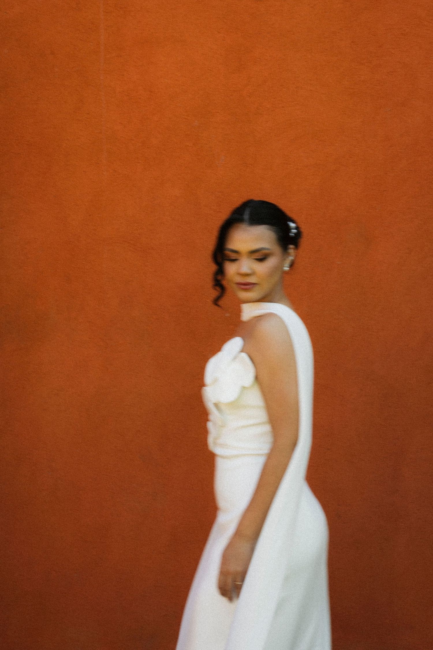 Bride in white gown with floral detail poses against vibrant orange wall.