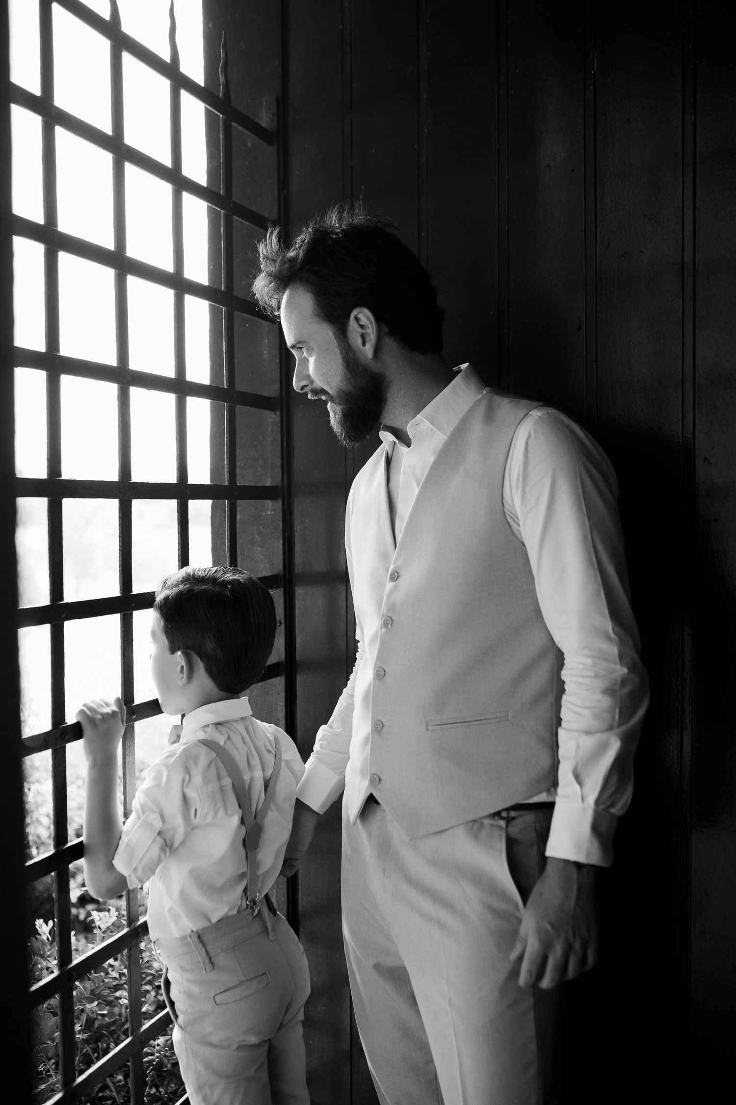Three figures in white formal attire look out large windows in dramatic black and white photo.