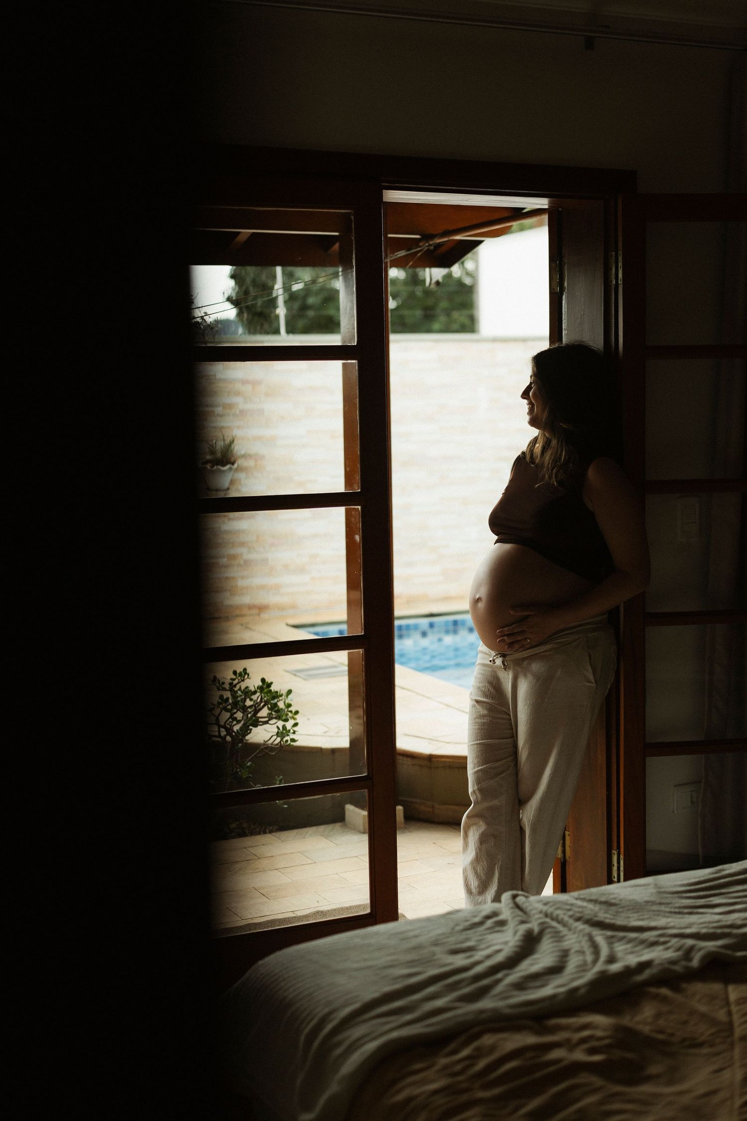 Pregnant woman standing in open doorway holding baby bump, backlit by natural light with pool view beyond.