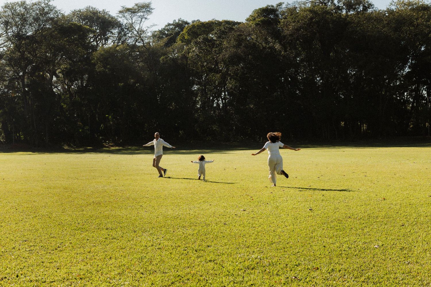 A joyful family with two adults and a child running and playing together on a lush green lawn surrounded by trees.
