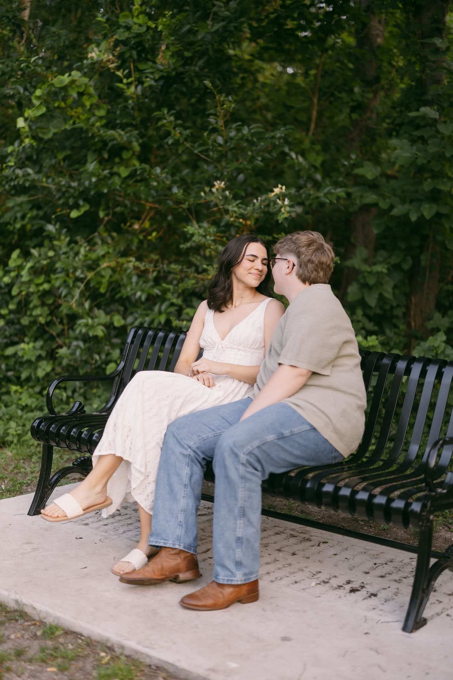 Couple sitting close together on a black park bench, surrounded by lush green trees, sharing an intimate moment.