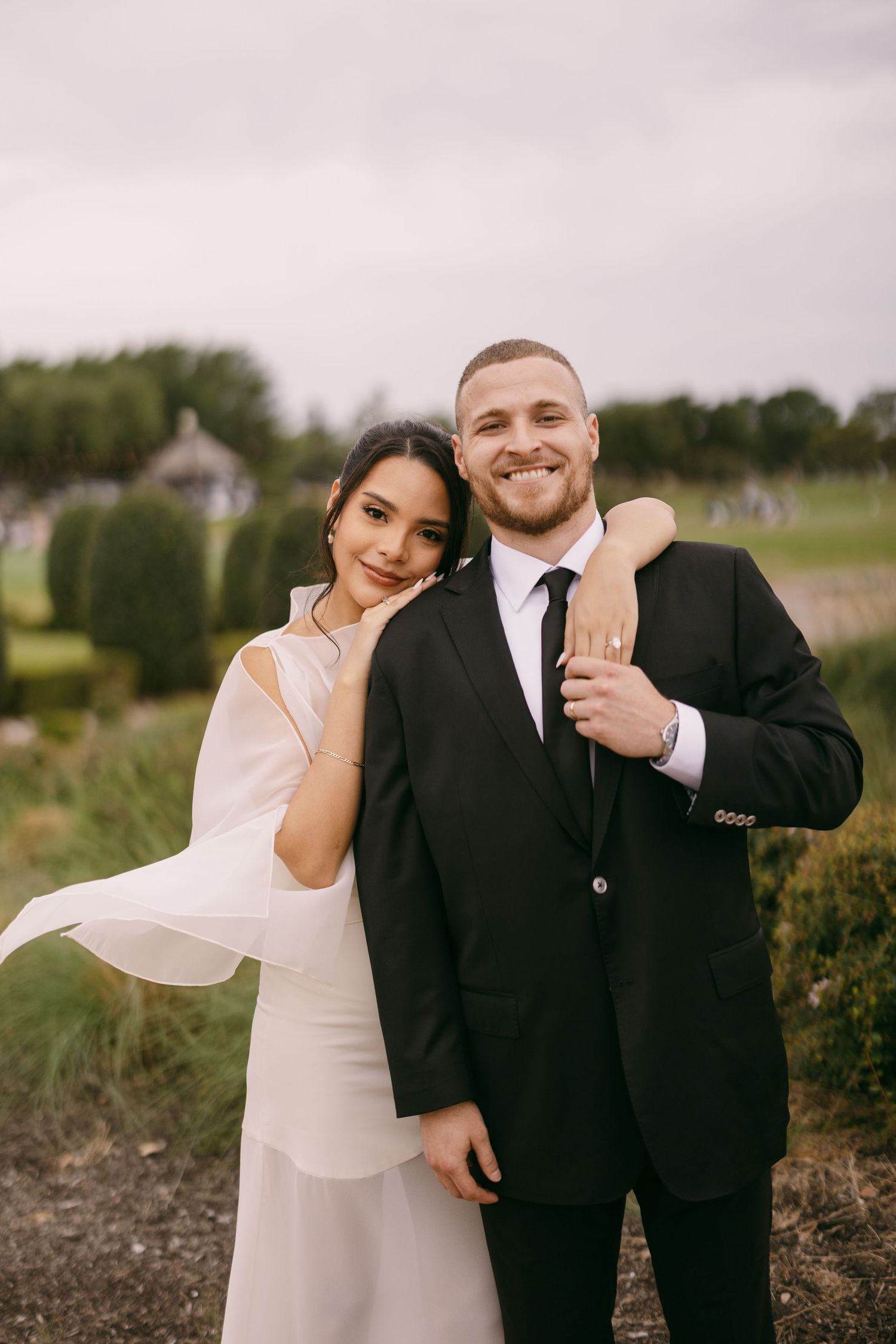 Happy newlyweds pose outdoors, bride in white gown with veil, groom in black suit, smiling at camera in garden setting.