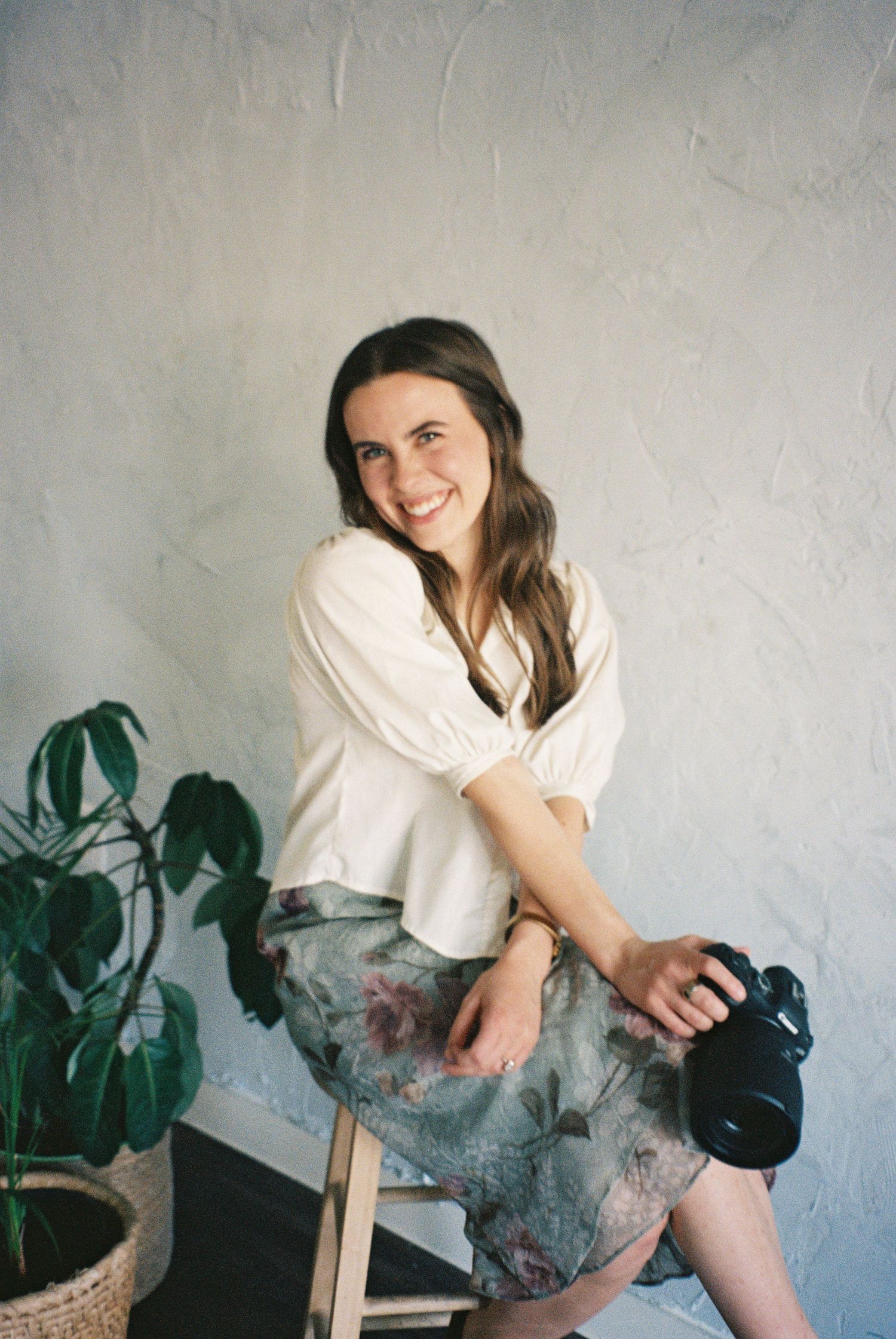 A woman in white blouse holds camera while sitting on stool beside houseplants.