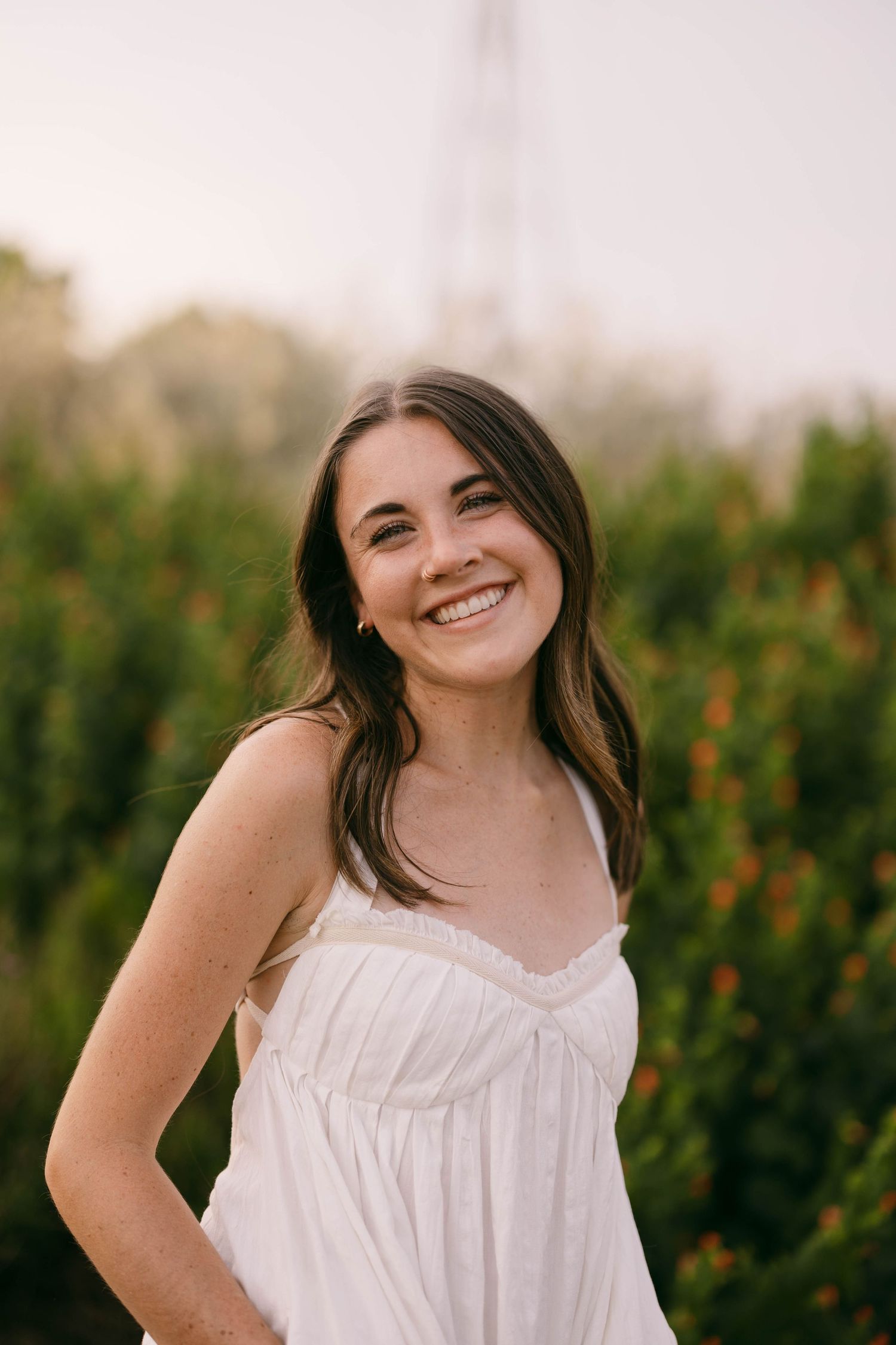 Person in white sleeveless top poses playfully against green foliage background during outdoor photoshoot.