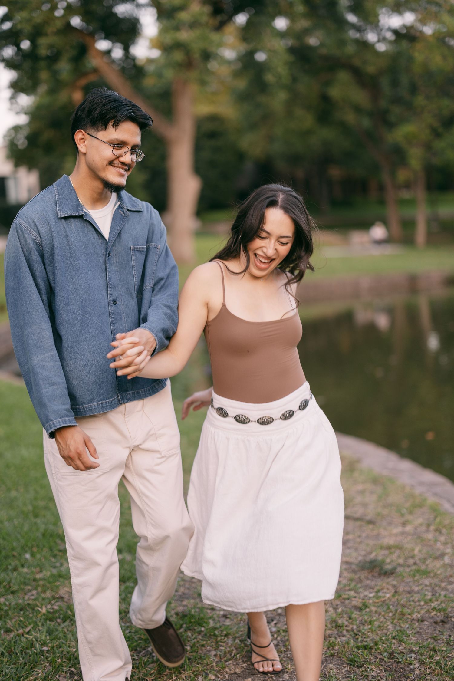 Happy couple walking hand in hand by a park pond, woman in brown top and white skirt, man in denim shirt.