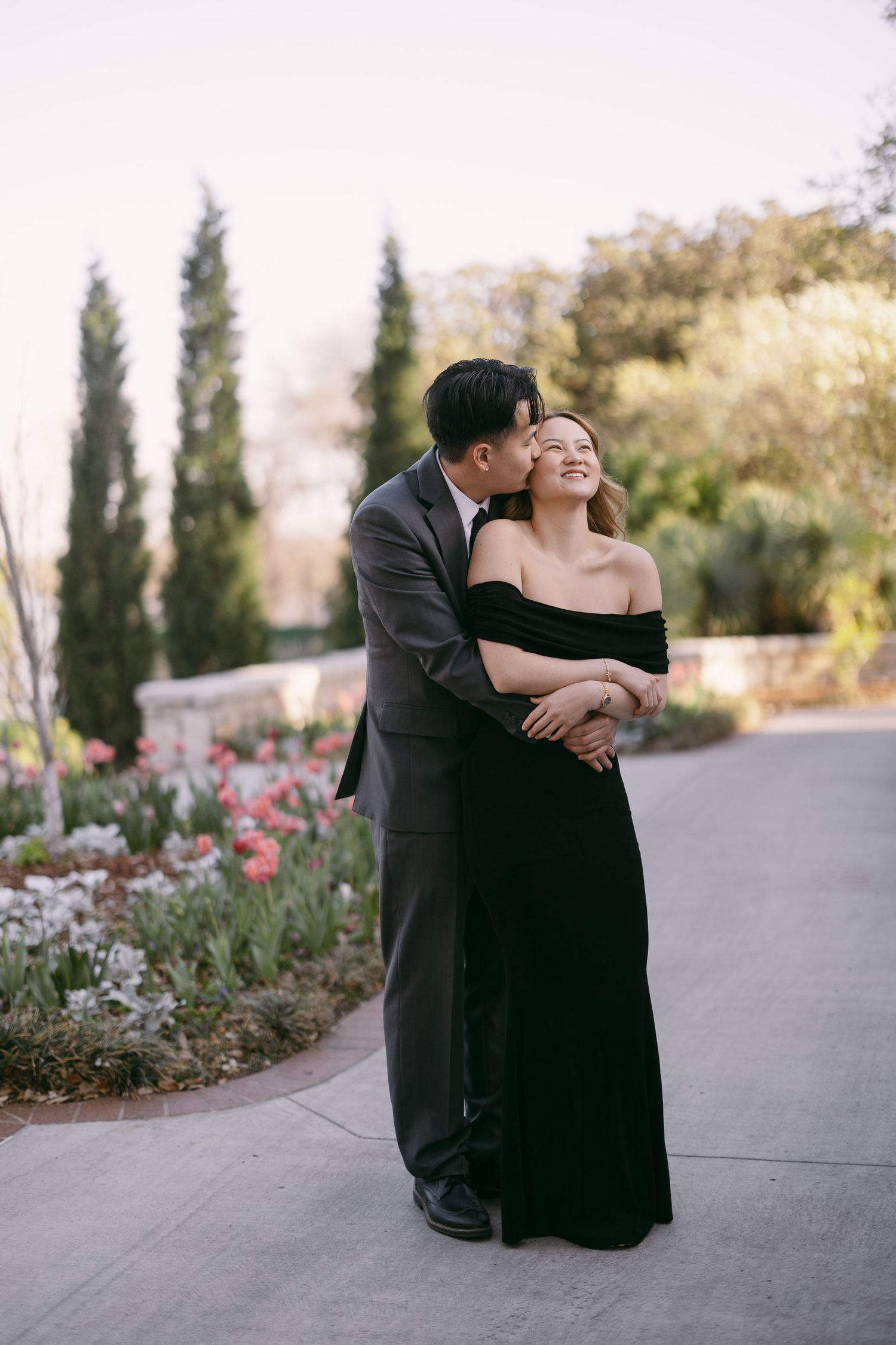 Couple embracing in garden, man kissing woman's cheek, she wears black off-shoulder gown, he wears gray suit, tulips in background.