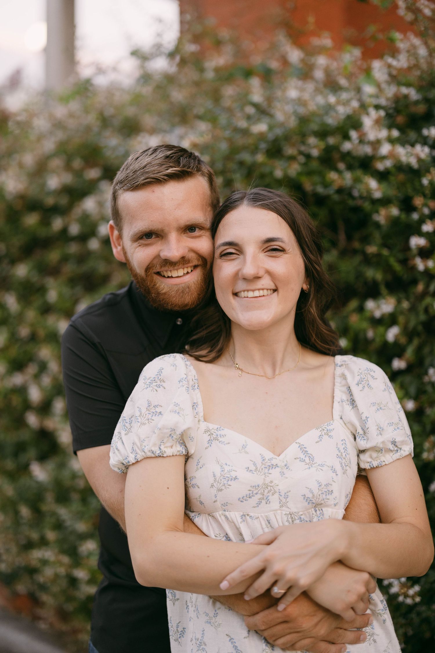 A couple embraces outdoors with a floral backdrop, wearing white and black casual attire.