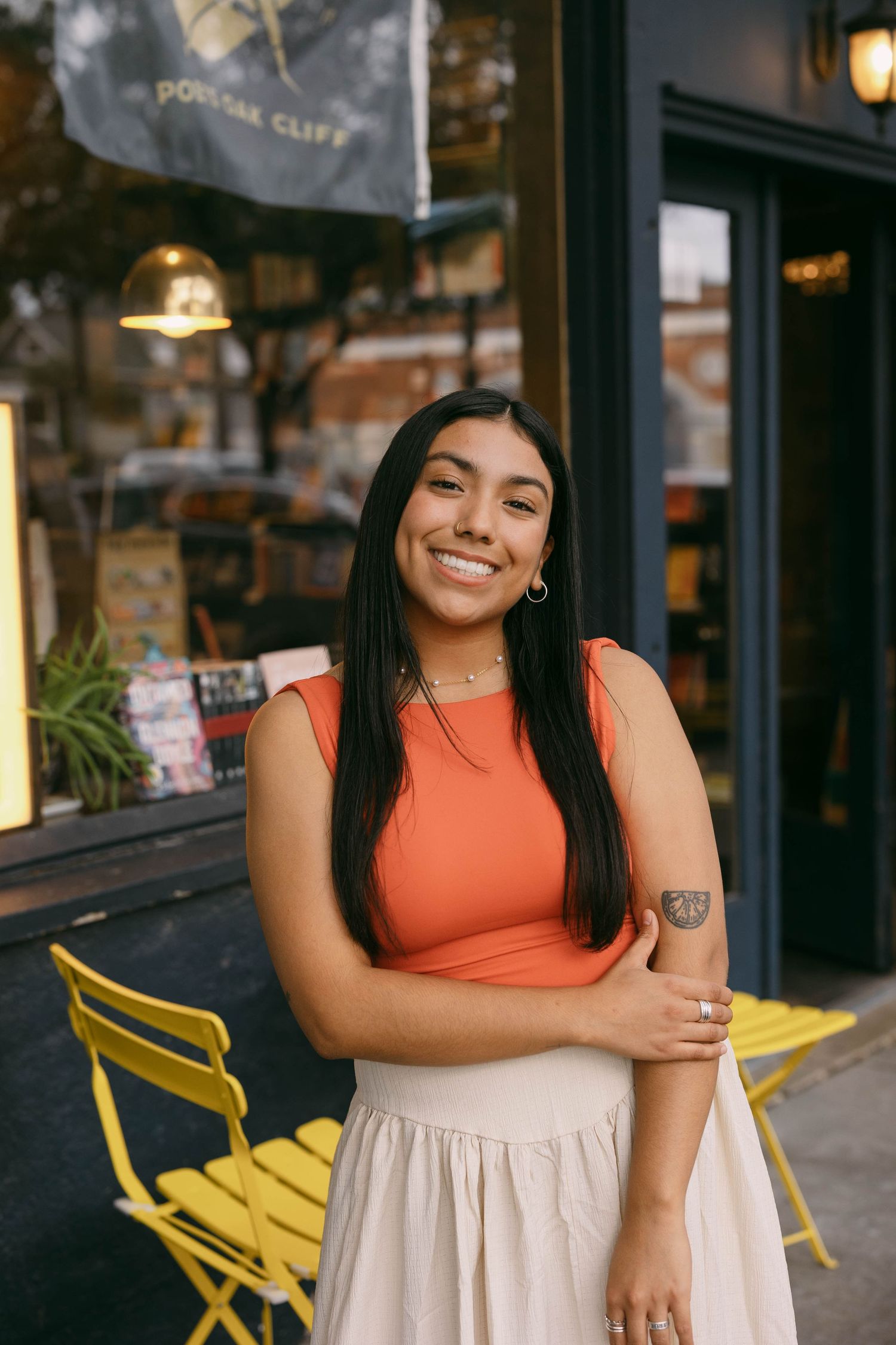 A person in an orange top and cream skirt poses casually against a storefront with yellow chairs.