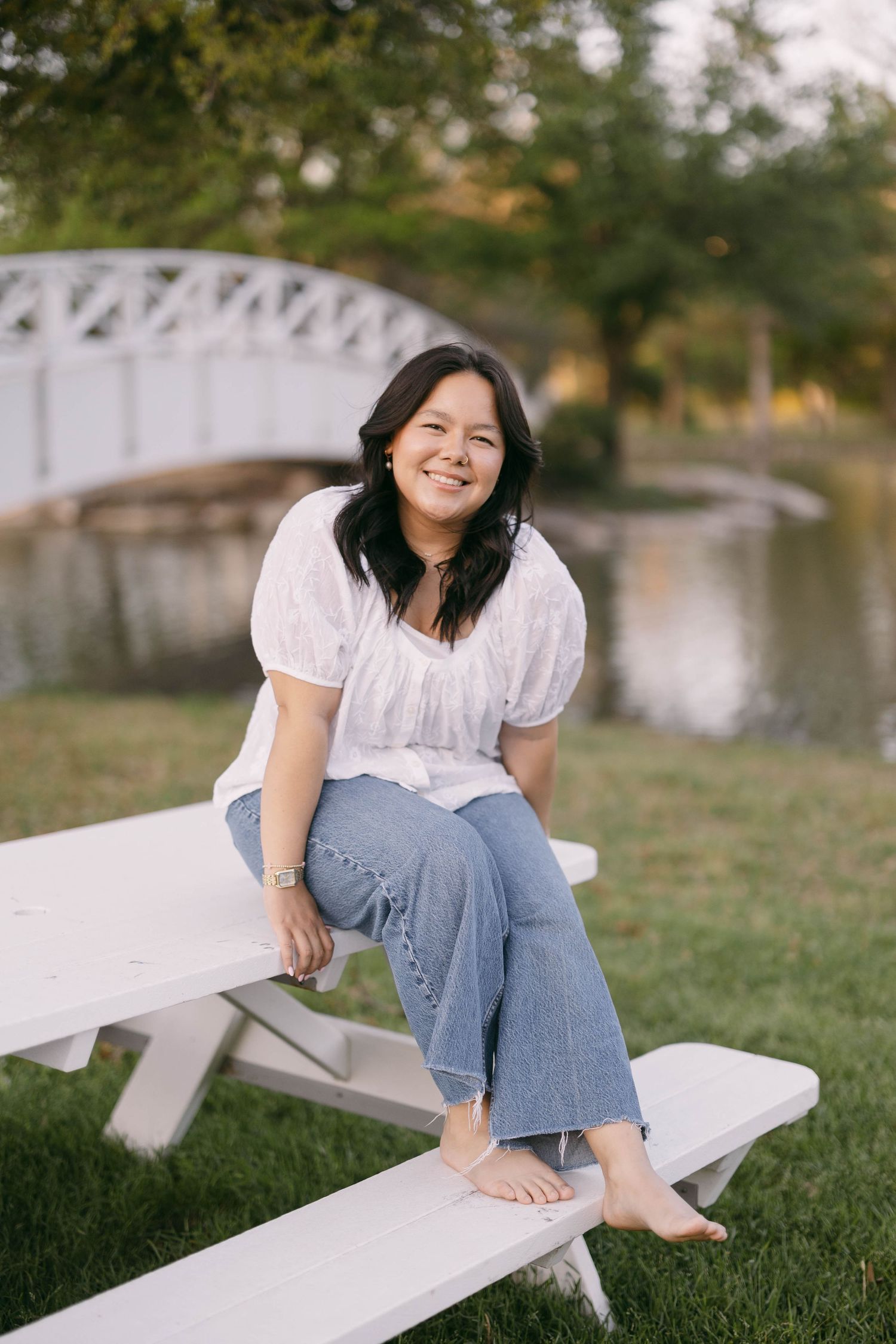 A woman in casual attire sits barefoot on a white bench near a scenic bridge and pond.