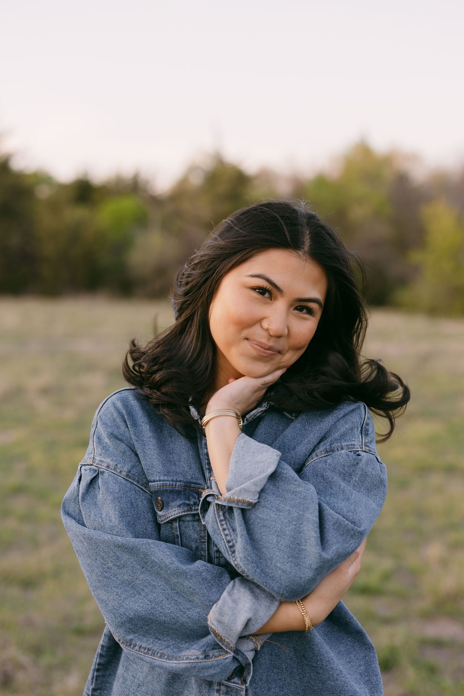 Young woman in denim jacket posing outdoors in a field at golden hour, smiling with hand on chin.