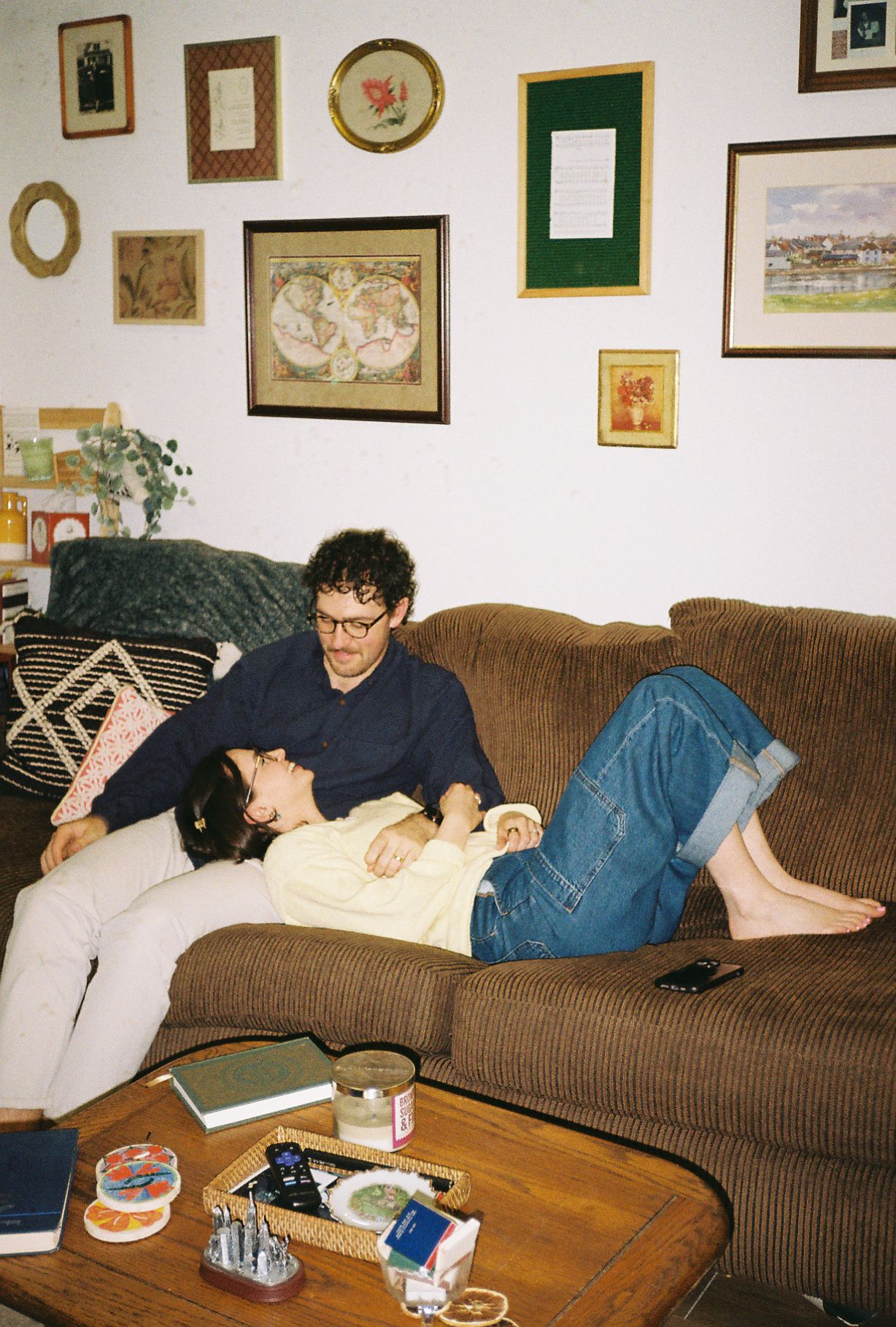 Couple relaxing on a brown corduroy couch surrounded by framed art and maps in a cozy, eclectic living room.