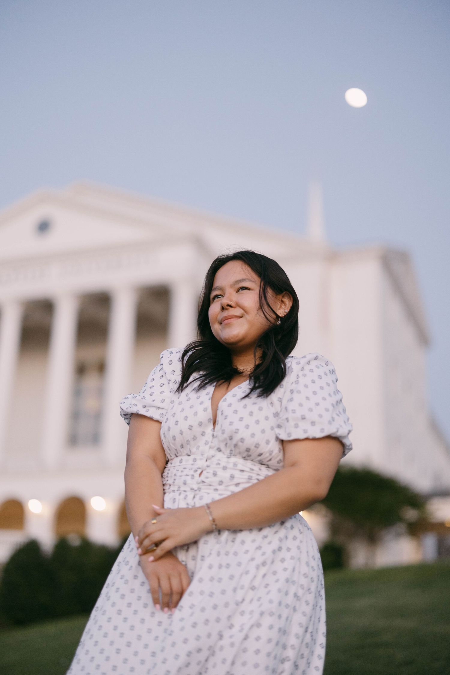 Person in polka dot dress poses on grassy hill with white building in background at dusk.