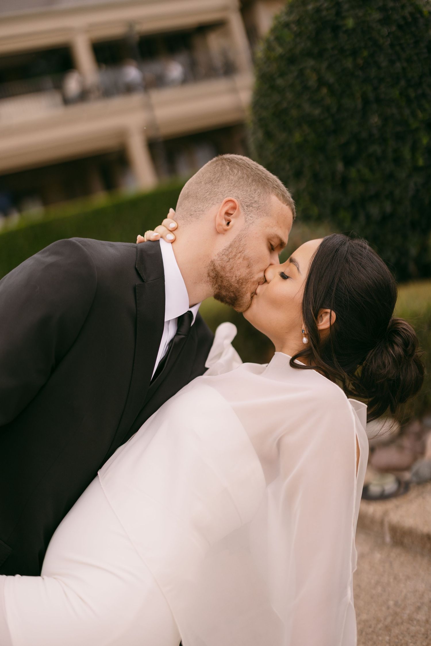 A bride and groom share a romantic kiss outdoors, with the groom in a black suit and bride in a white gown.