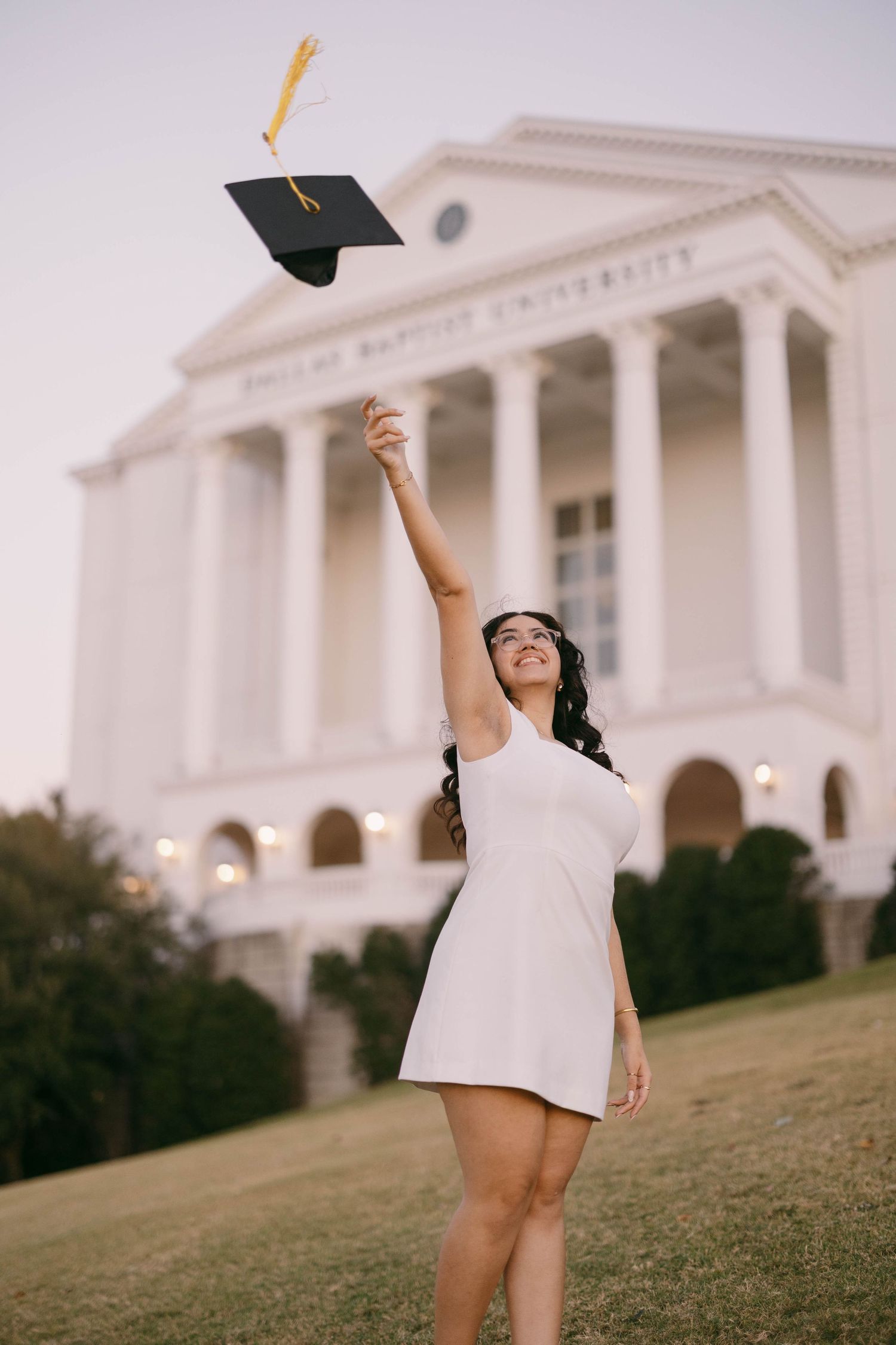 Person in white dress throws graduation cap in front of classical white building.
