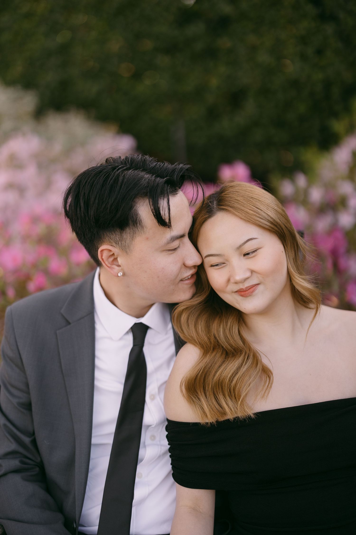 A couple shares a tender moment, man in suit kissing woman's cheek near pink flowers.
