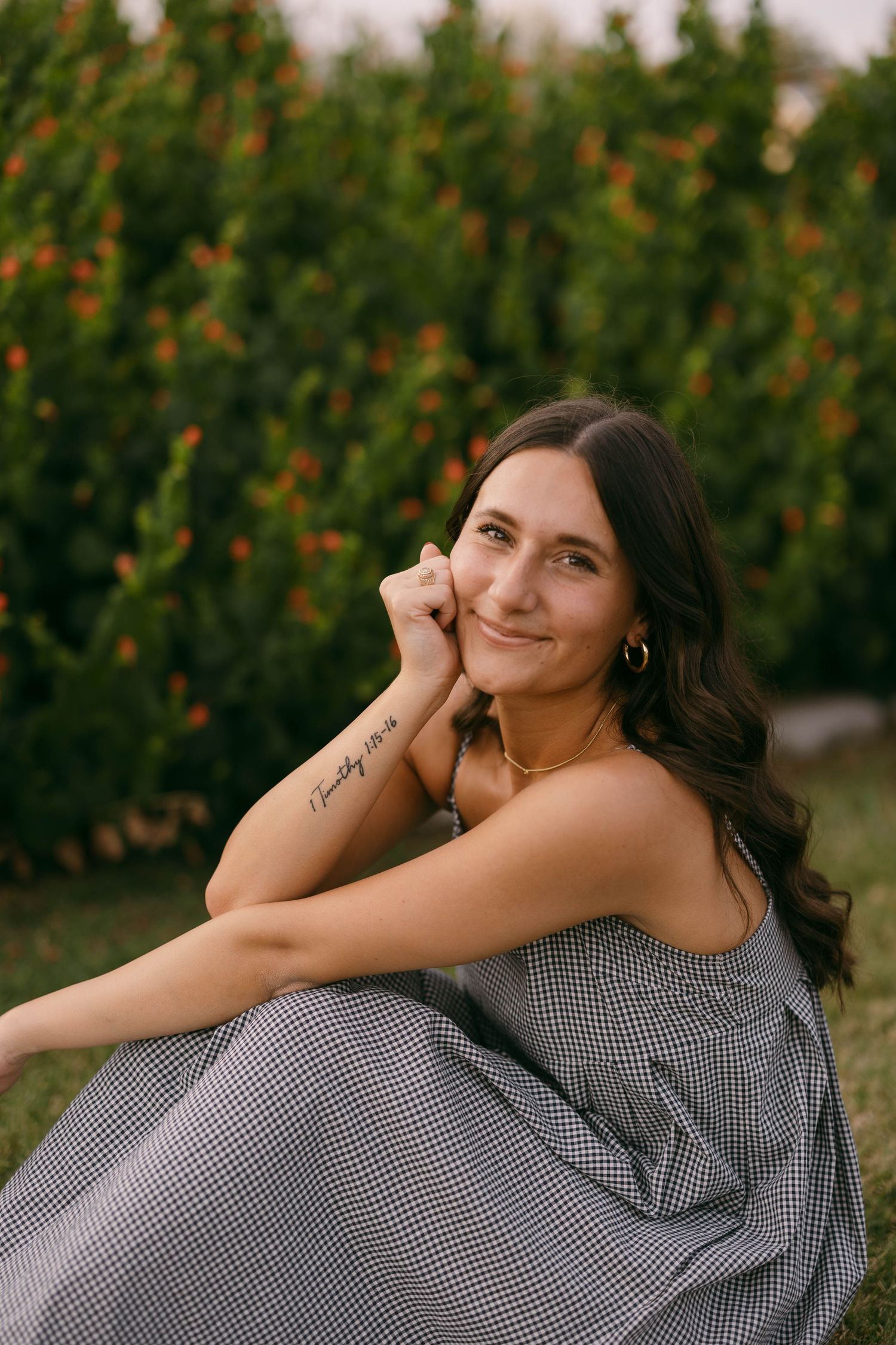 A woman in gingham dress poses outdoors against flowering bushes at sunset.