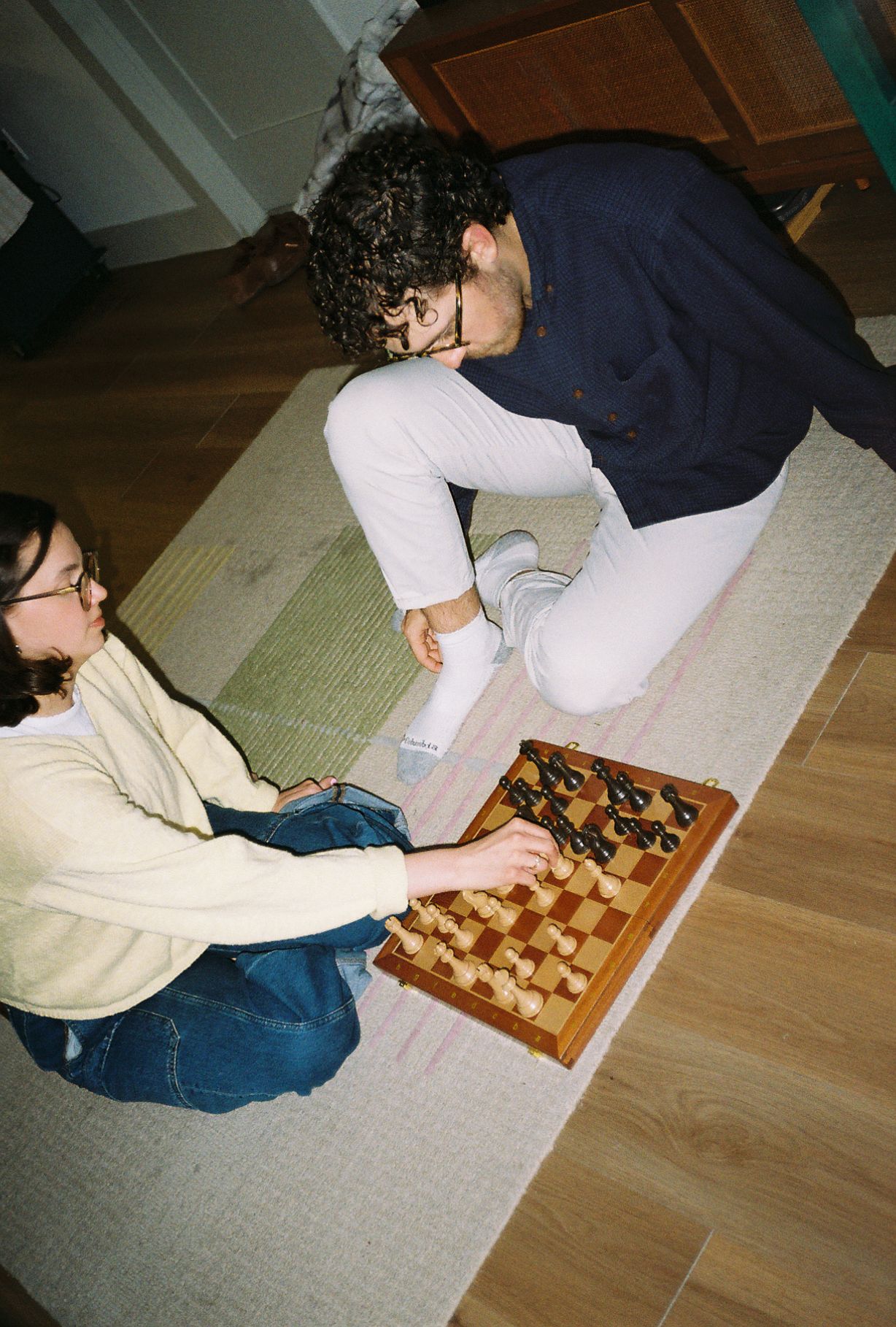 Two people playing chess on a wooden board while sitting on a rug on the floor indoors.
