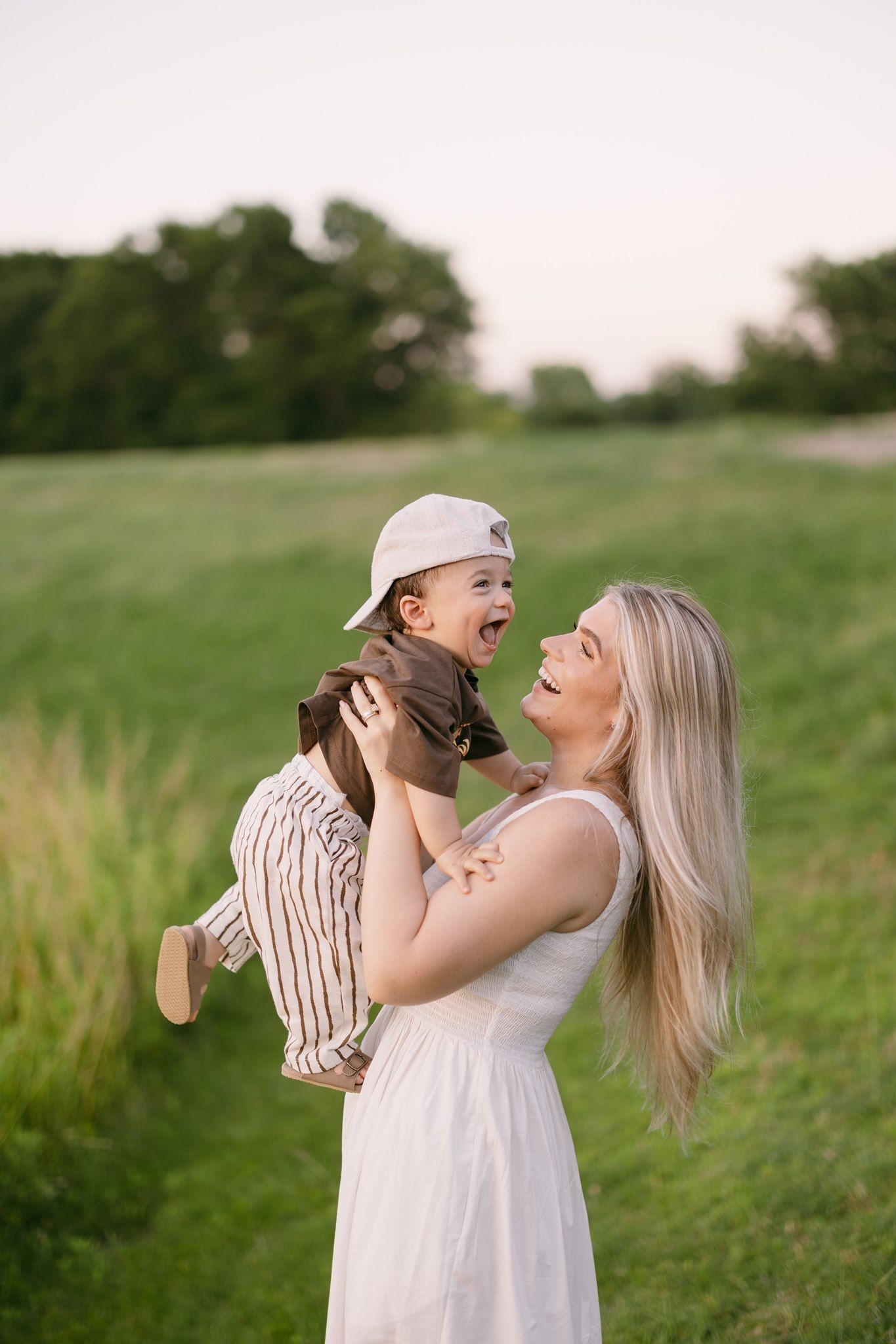 A joyful blonde woman in a white dress lifts a laughing toddler in a cap outdoors on a green field.