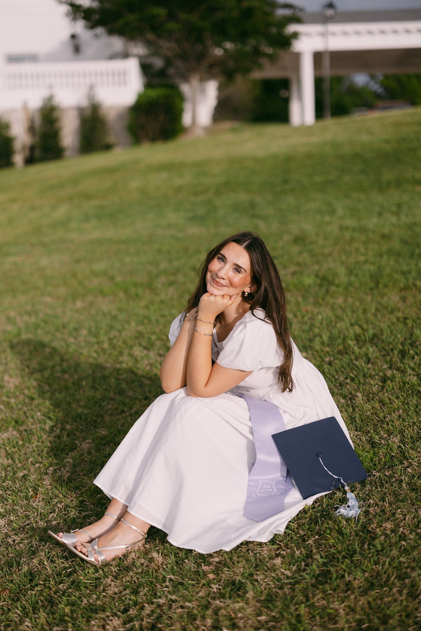 Young woman in white dress sitting on grass with graduation cap, smiling at camera during outdoor graduation photo session.