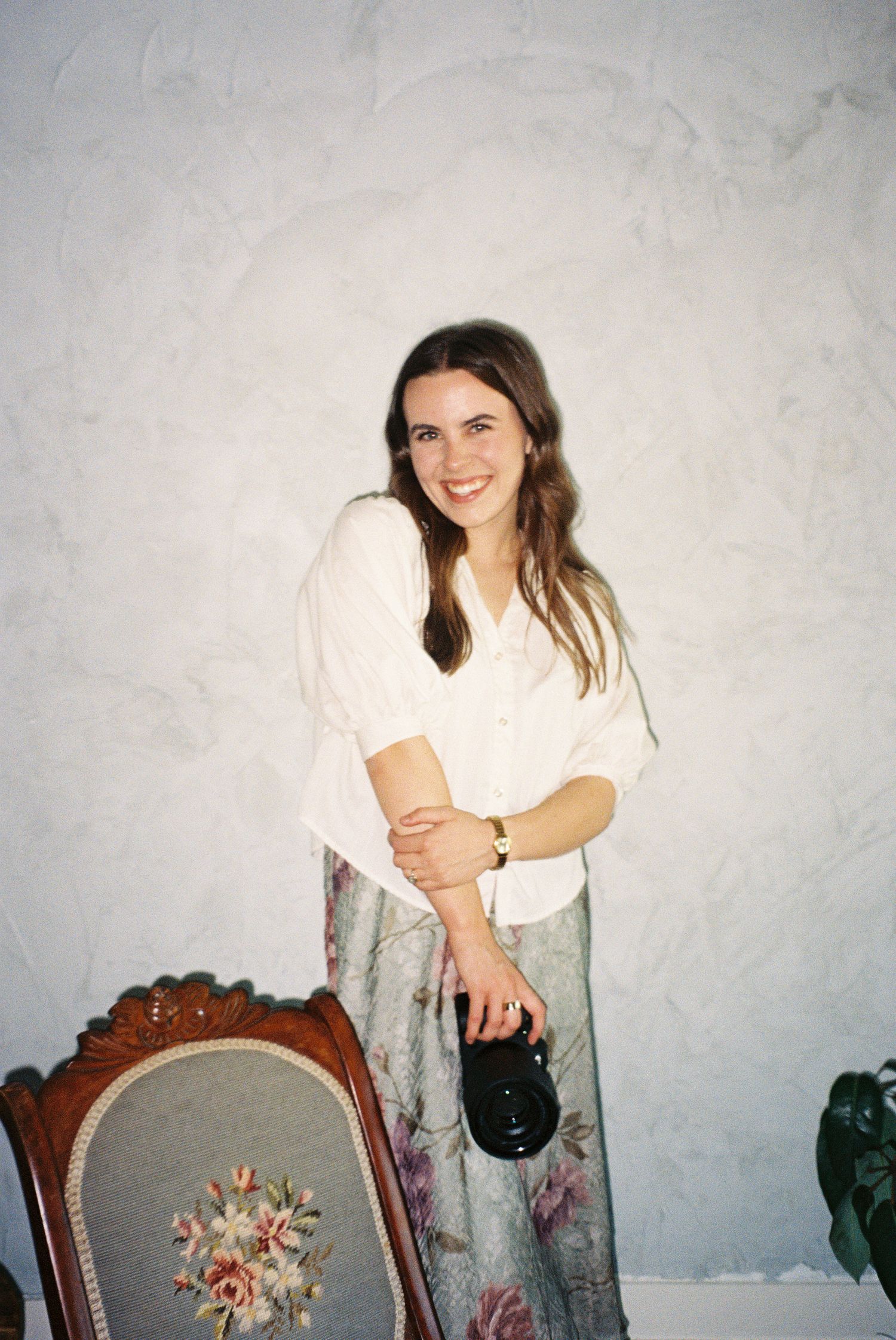 A woman in a white blouse and floral pants poses next to an antique chair with embroidered upholstery.