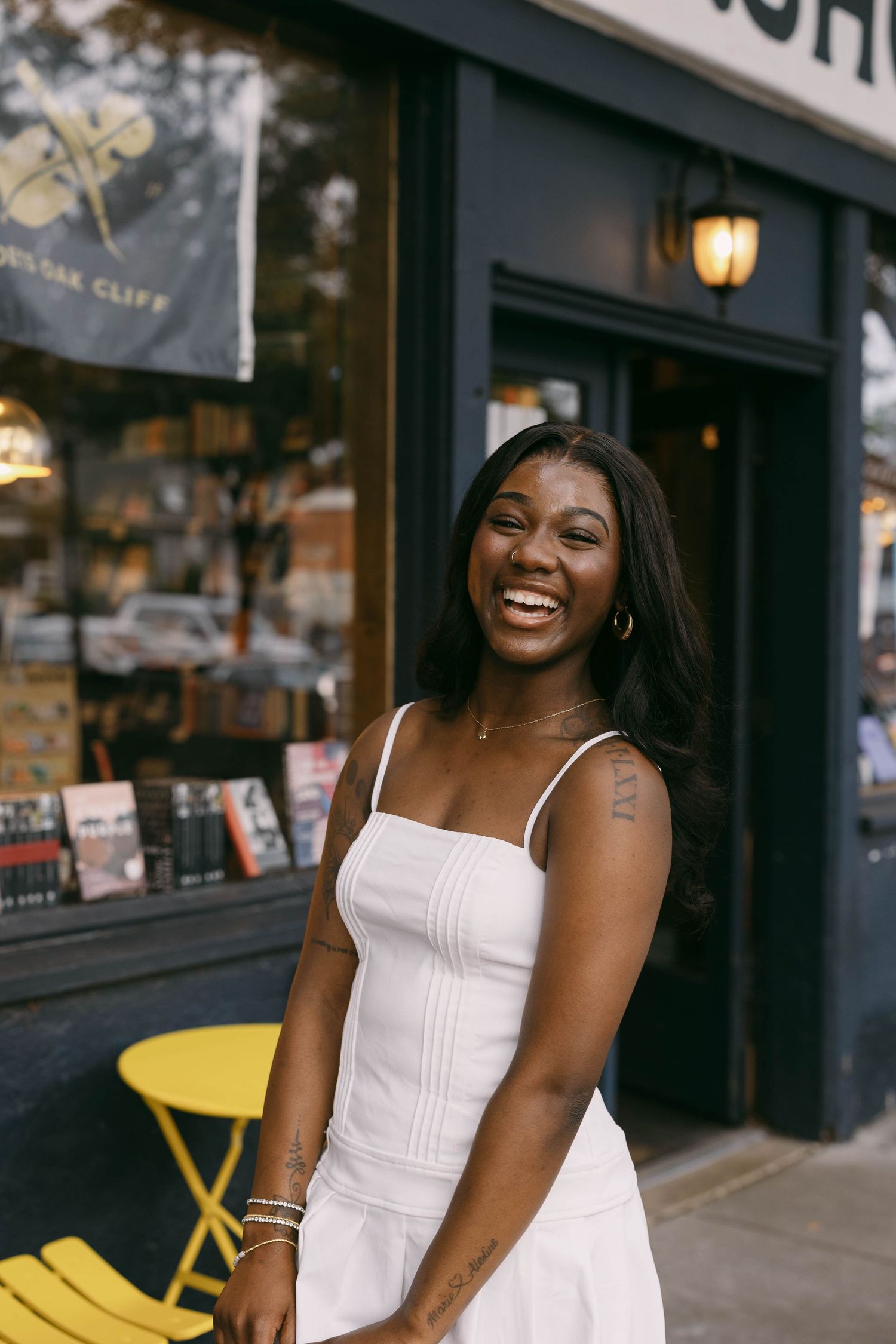 Person in white dress poses cheerfully outside storefront with yellow chairs.