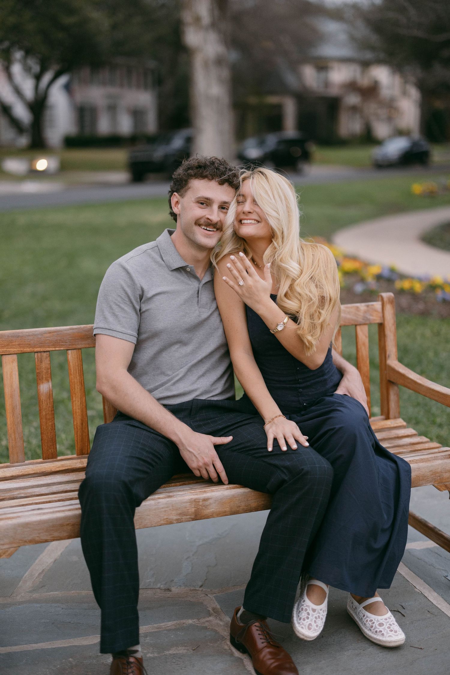 Happy couple sitting on a wooden bench outdoors, woman showing engagement ring, smiling together in a park setting.