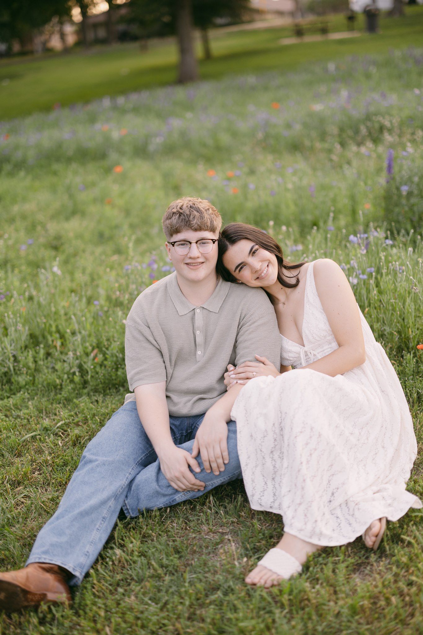 A young couple sits together in a wildflower meadow, smiling, as she leans on his shoulder in a white lace dress.