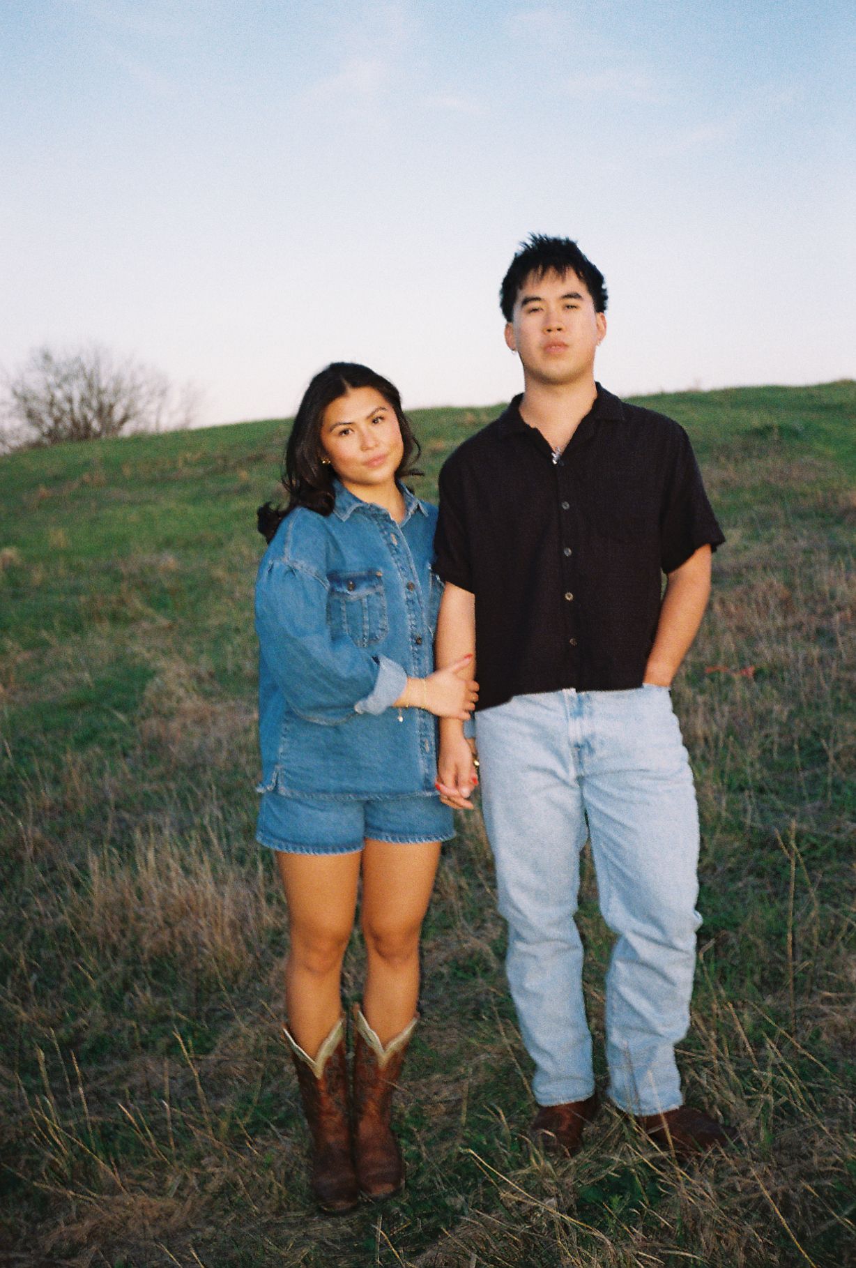 A young couple holds hands in a grassy field, woman in denim romper and cowboy boots, man in black shirt and jeans.