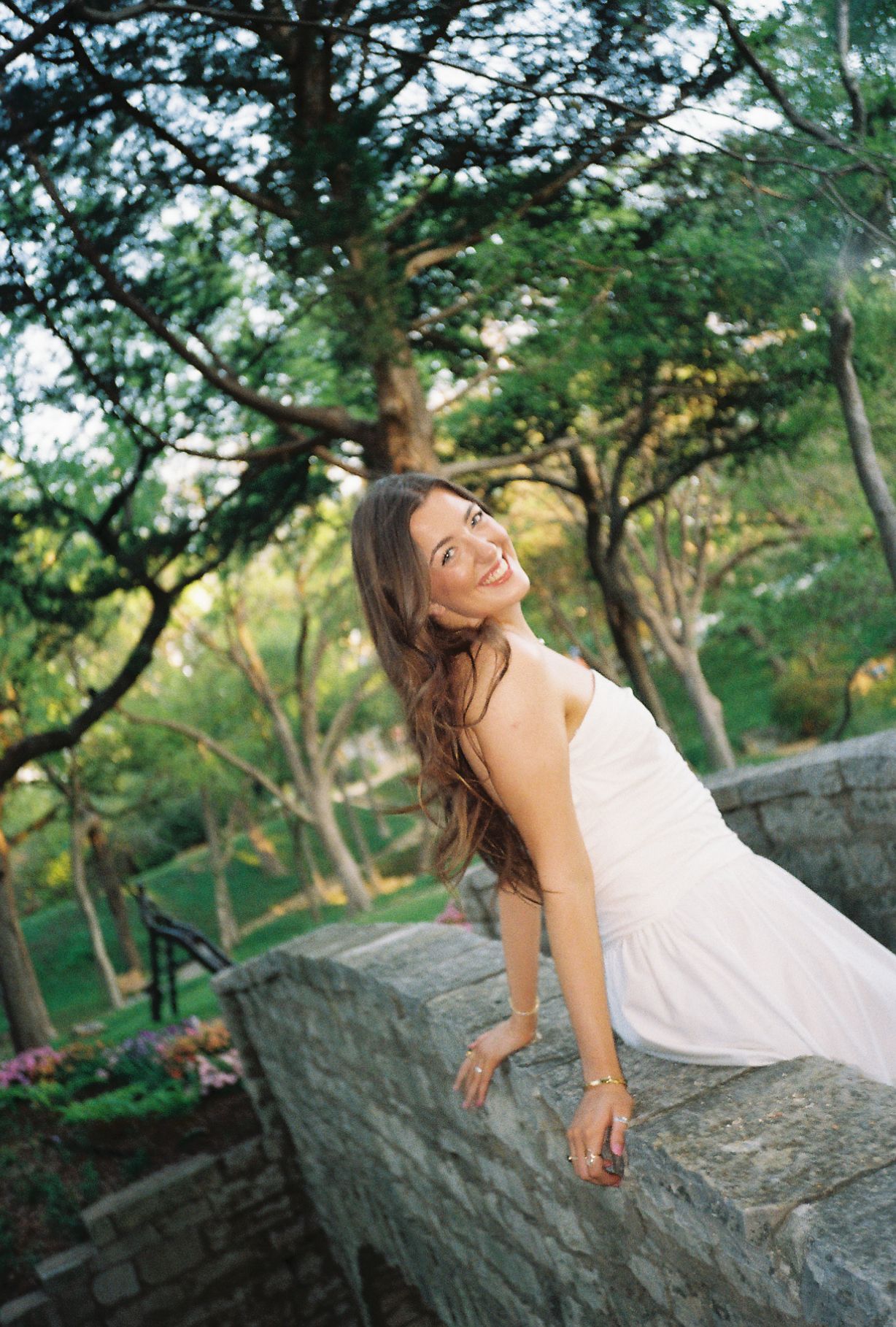 A smiling young woman in a white dress leans on a stone wall surrounded by lush green trees in a garden.