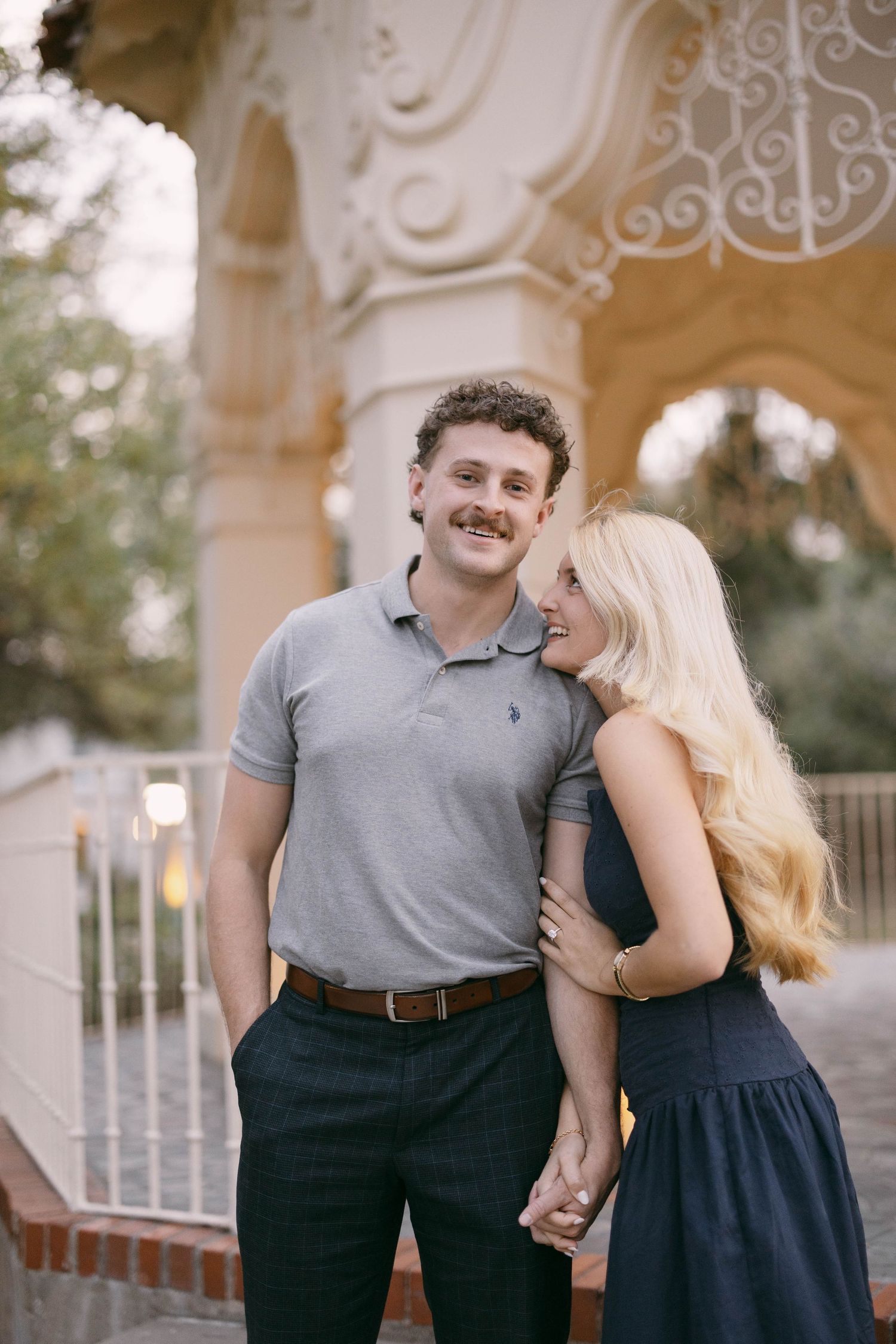 A smiling couple holds hands outdoors near ornate architecture, man in gray polo and woman in navy dress.
