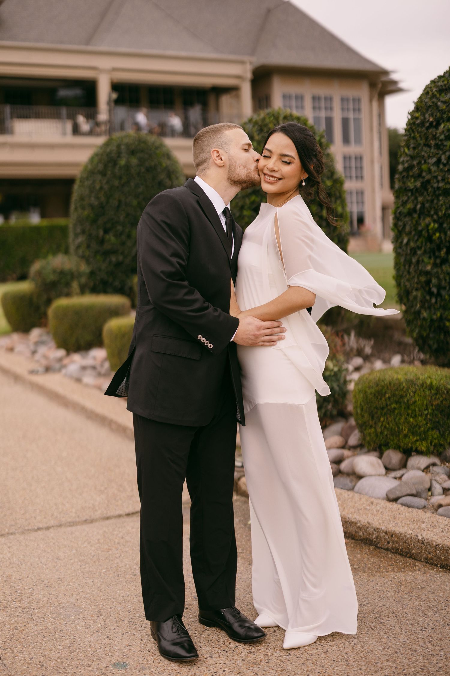 Couple in wedding attire embrace outdoors, groom in black suit kisses bride in flowing white dress near manicured hedges.
