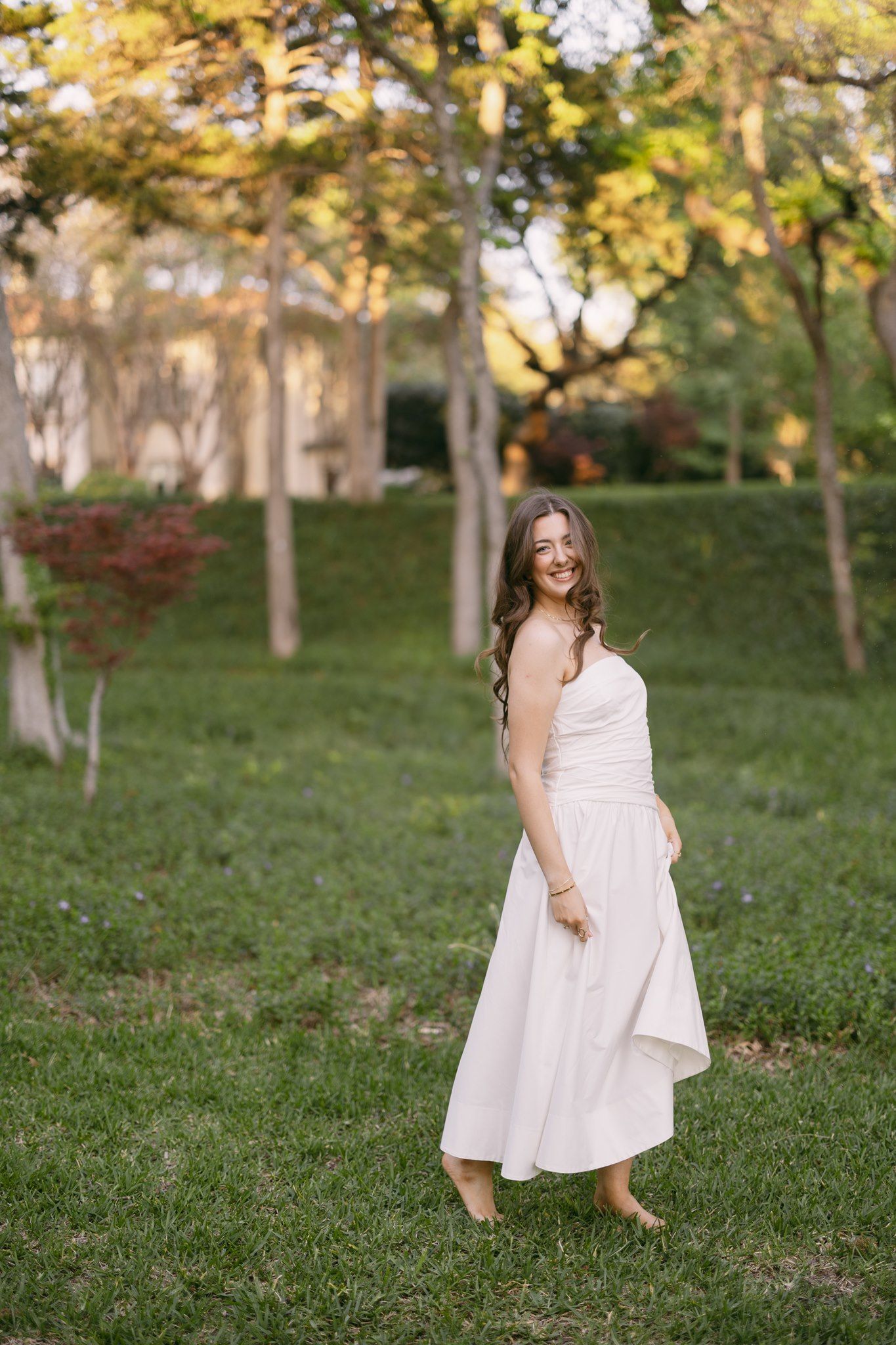 Smiling woman in white strapless midi dress standing barefoot on green grass in a sunlit park.