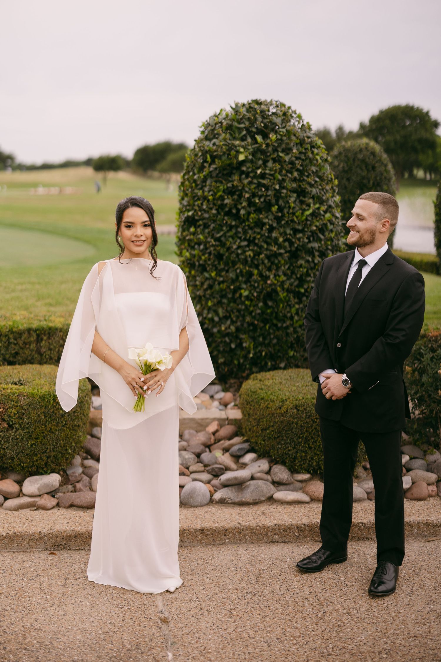 Bride in white cape dress holding bouquet stands beside groom in black suit at outdoor garden wedding venue.