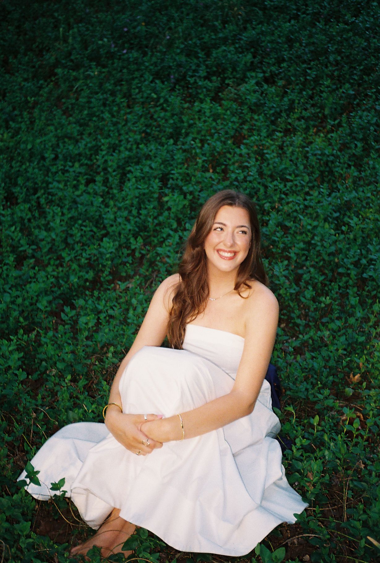 Pregnant woman in white strapless dress sitting on green ivy, smiling warmly during outdoor maternity photo session.