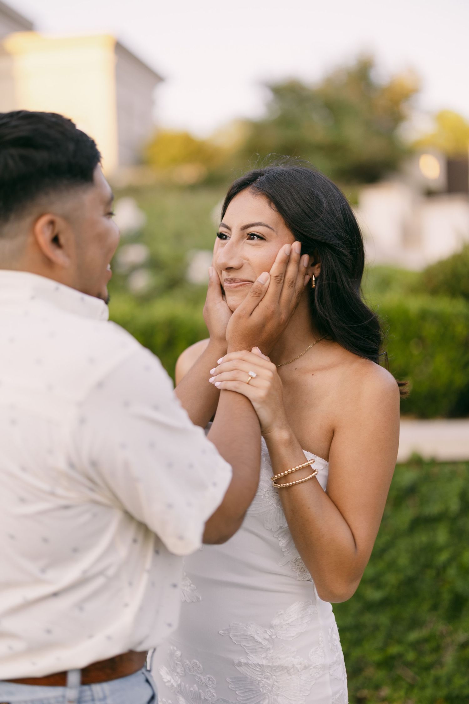 A couple shares a tender moment outdoors, man gently cupping womans face, she wears white dress and engagement ring.