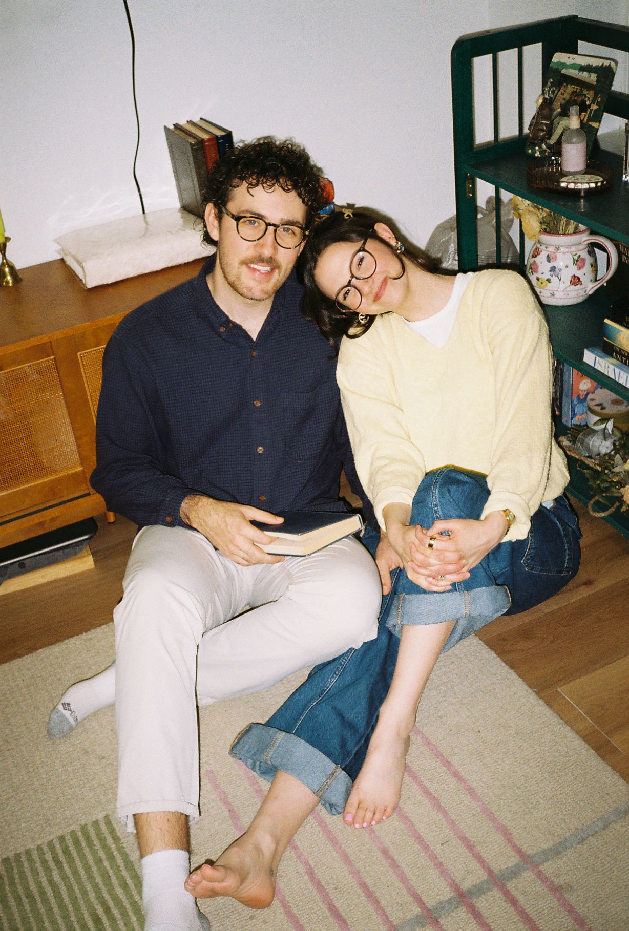 A couple wearing glasses sits together on a rug, smiling warmly, one in navy shirt and one in yellow sweater.