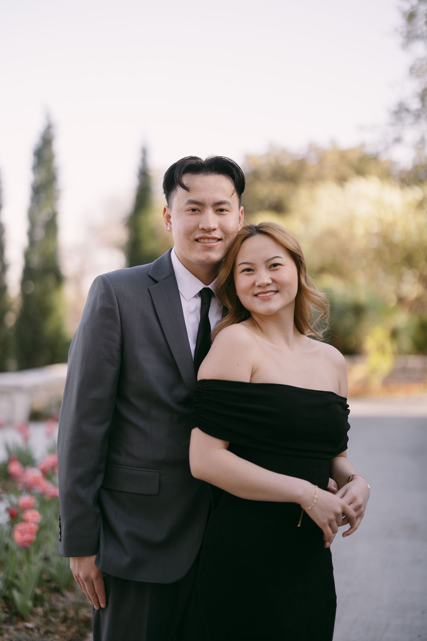 Couple in formal attire posing outdoors, man in gray suit and woman in black off-shoulder dress, smiling together.