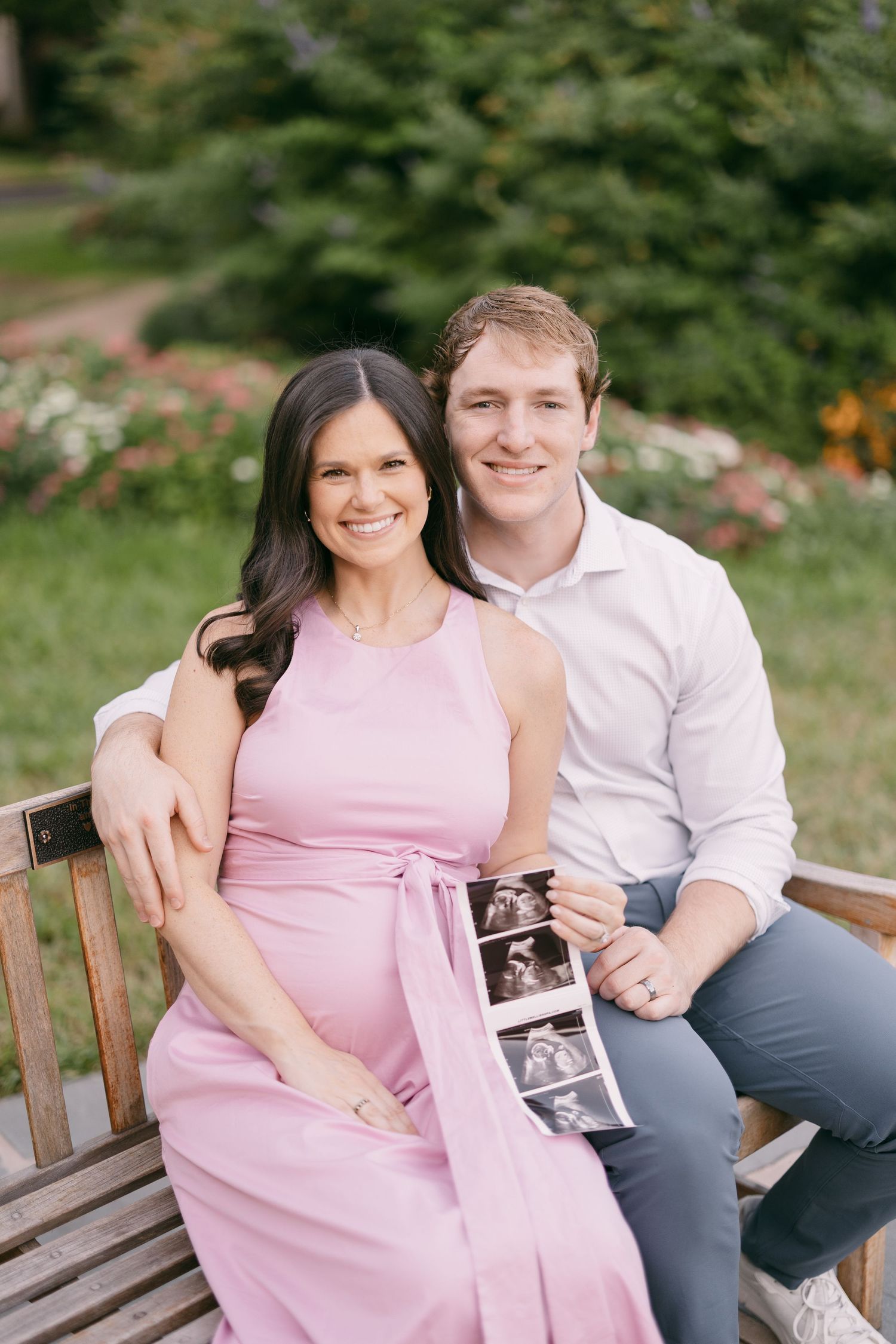 Smiling pregnant couple sitting on a wooden bench outdoors, holding ultrasound photos, surrounded by lush greenery.