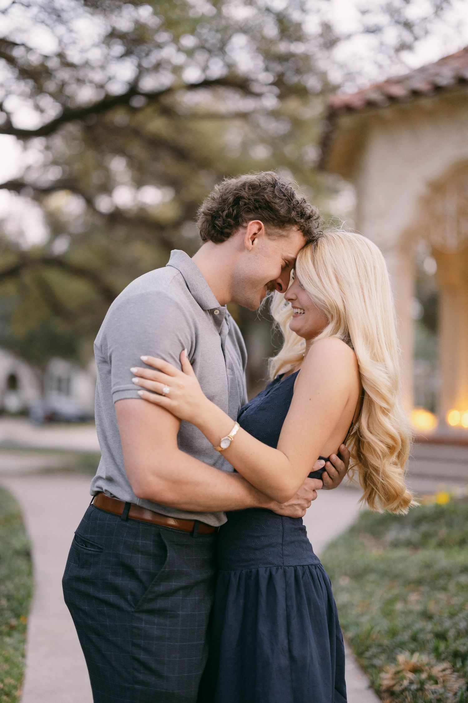 Couple embracing outdoors near a building, man in gray polo and woman in navy dress sharing a romantic moment.