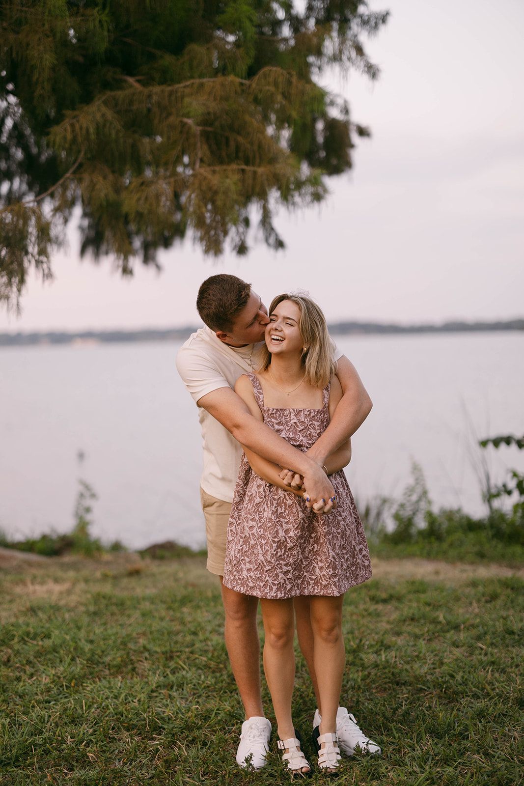 Couple in casual outfits embracing by a lakeside during sunset with trees in the background.