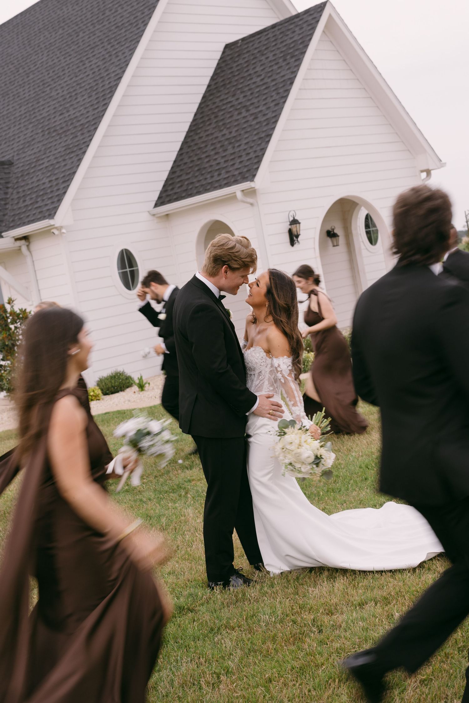 Bride and groom share a kiss outside a white chapel surrounded by wedding party in brown dresses and black tuxedos.