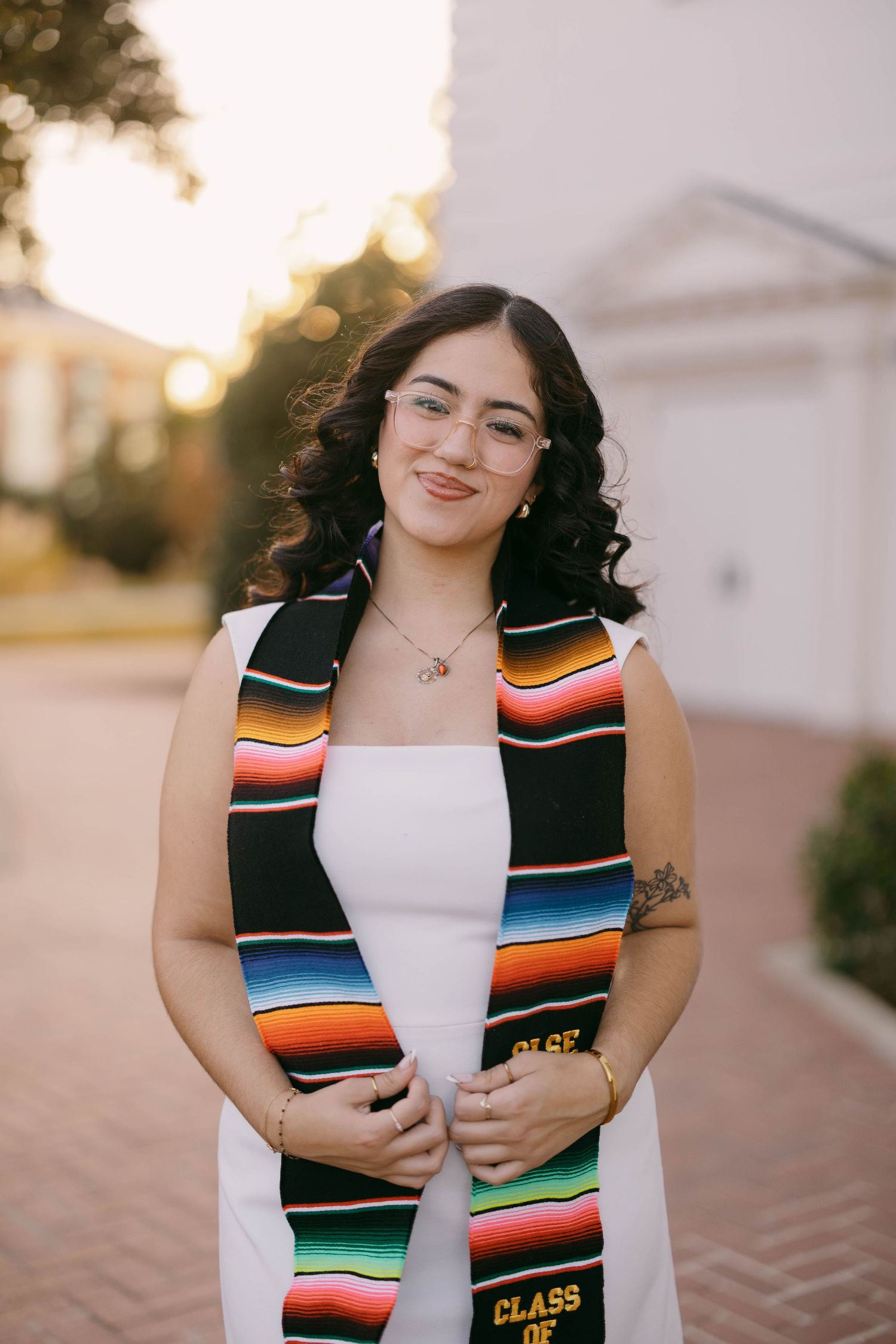 Graduate wearing a colorful serape graduation stole and white dress poses outdoors.