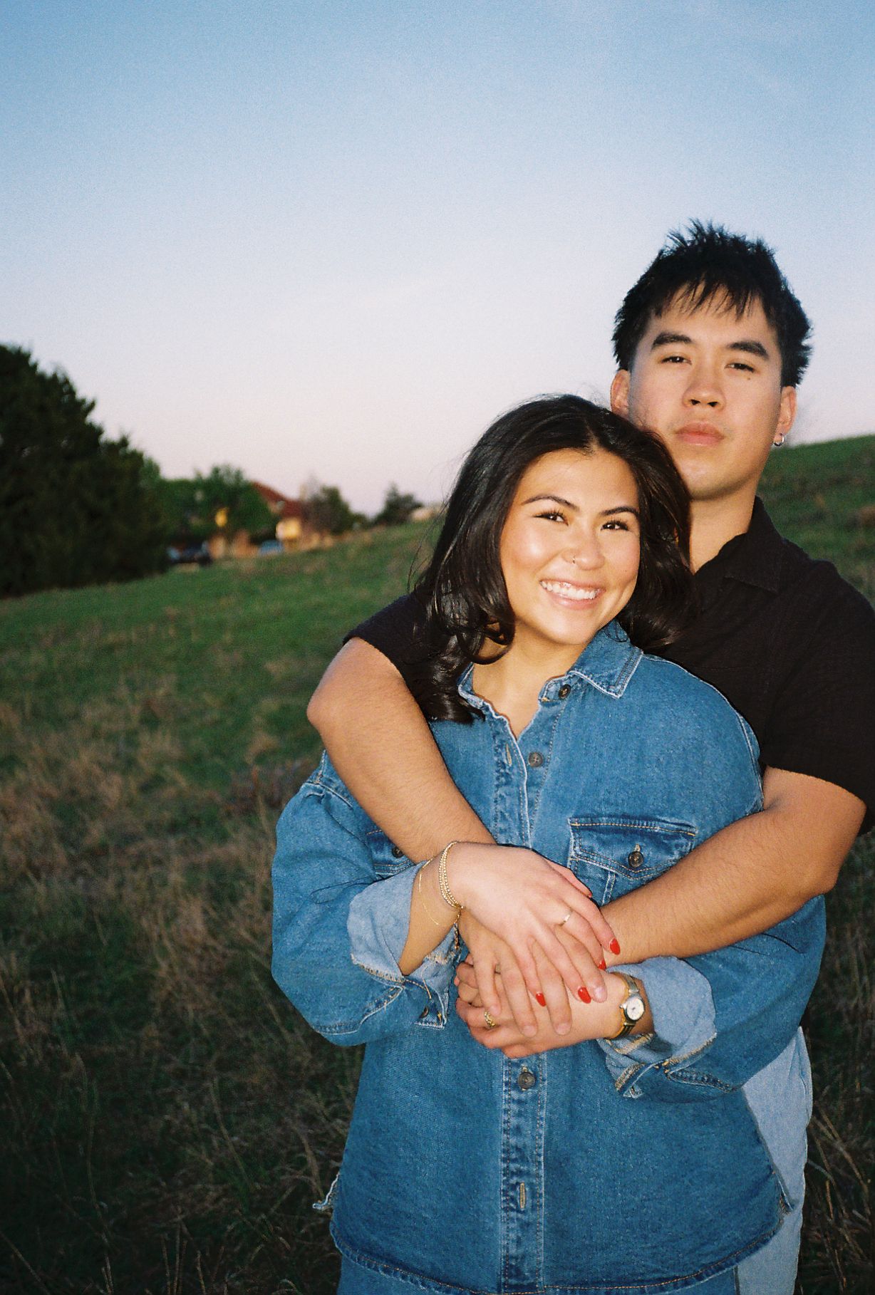 A smiling couple embraces outdoors on a green hillside at dusk, woman in denim jacket with man in black shirt behind her.