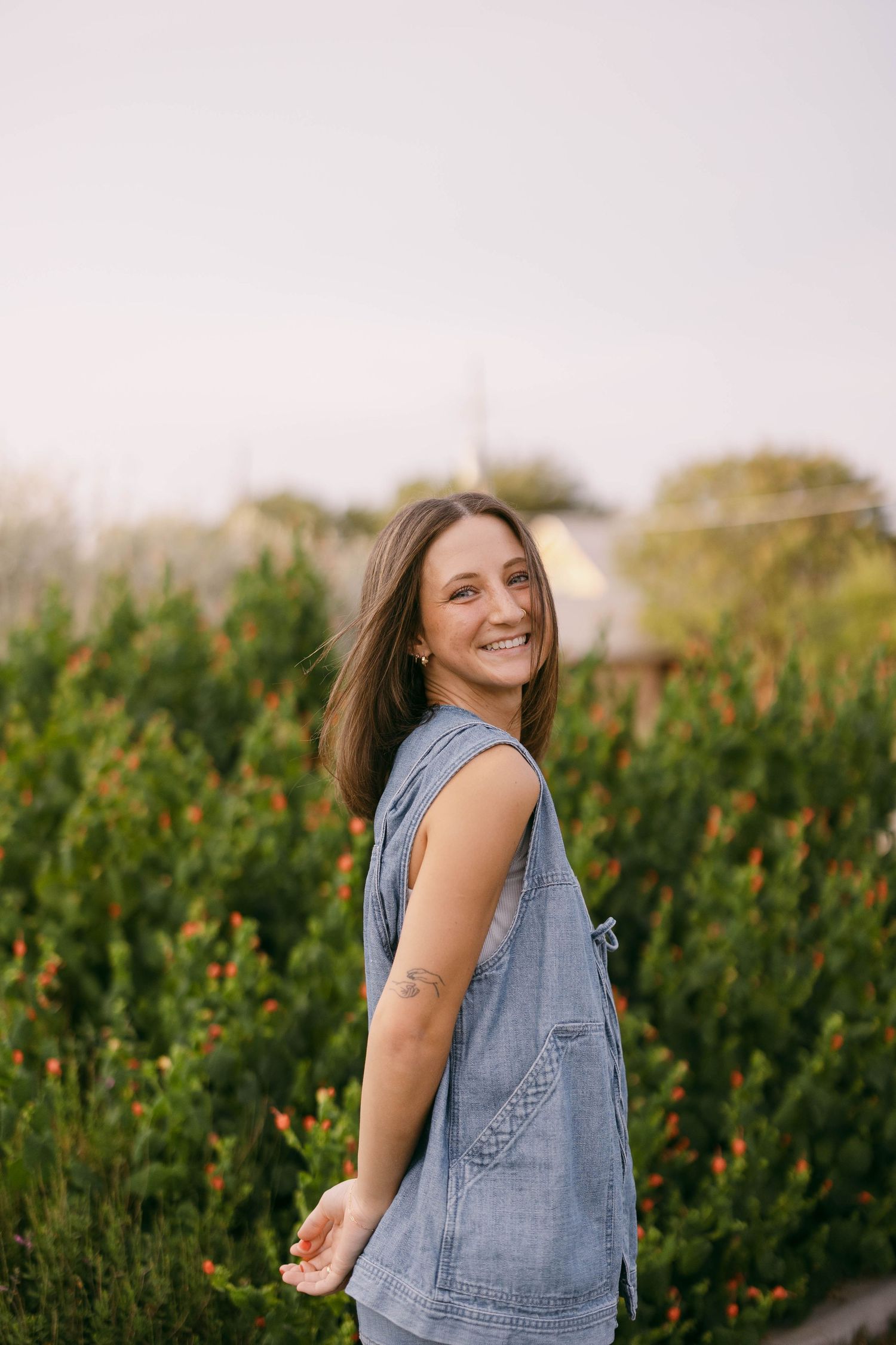 Person in denim overalls dances joyfully against green foliage during sunset photoshoot.