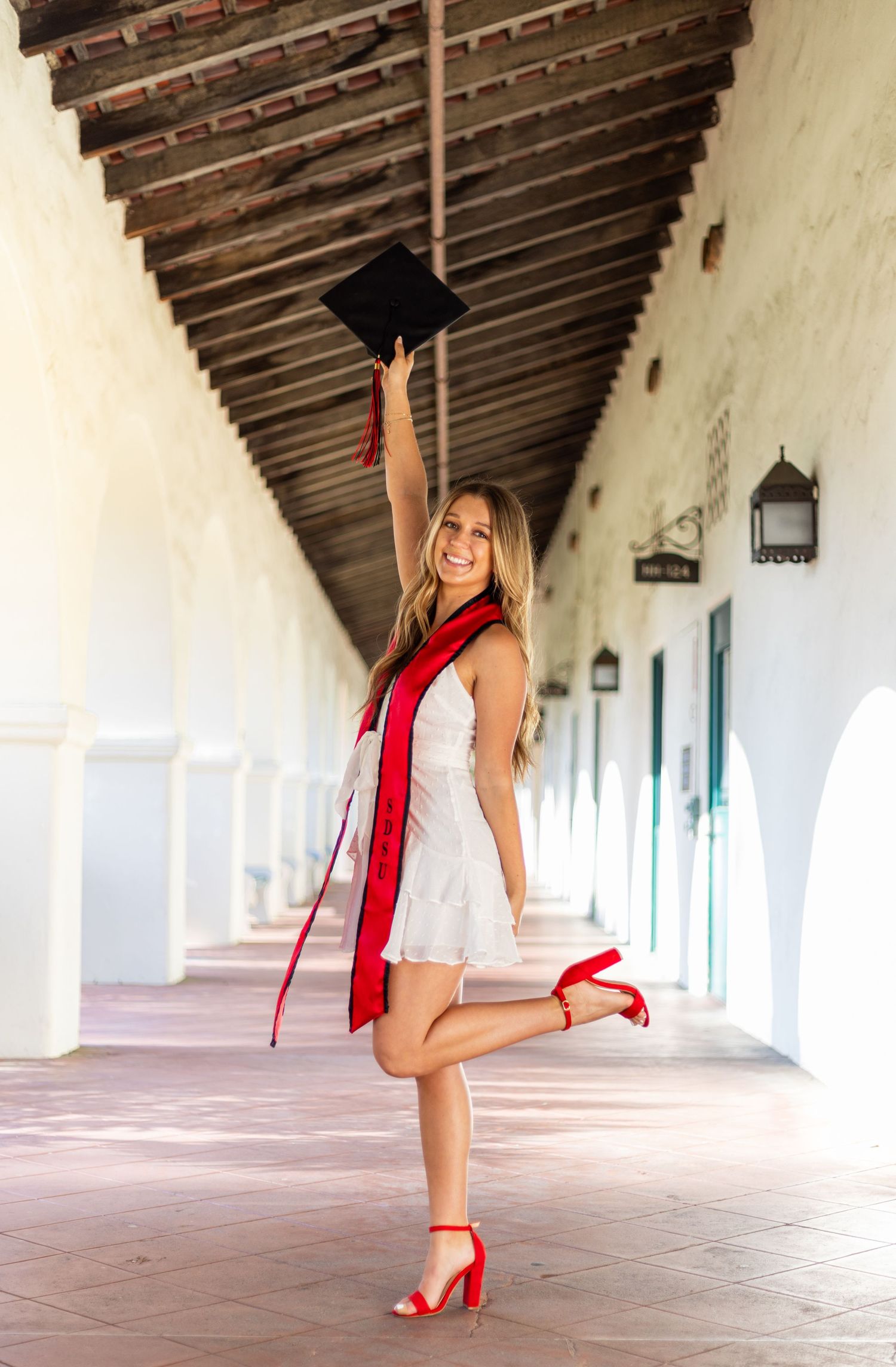 Graduate in white dress and red stole poses playfully in historic colonnade.