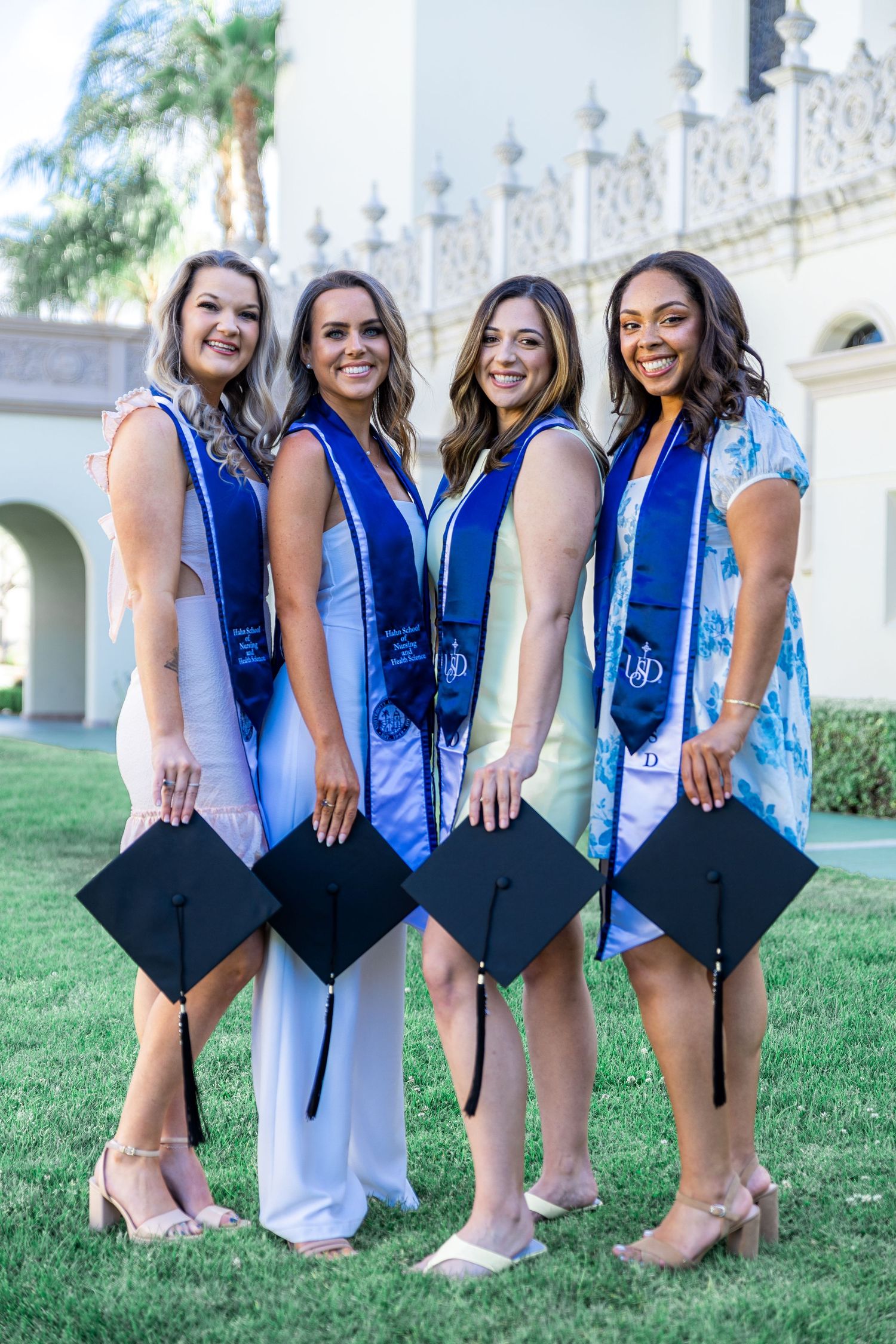 Graduates display their mortarboards while posing on grass in front of white mission building.