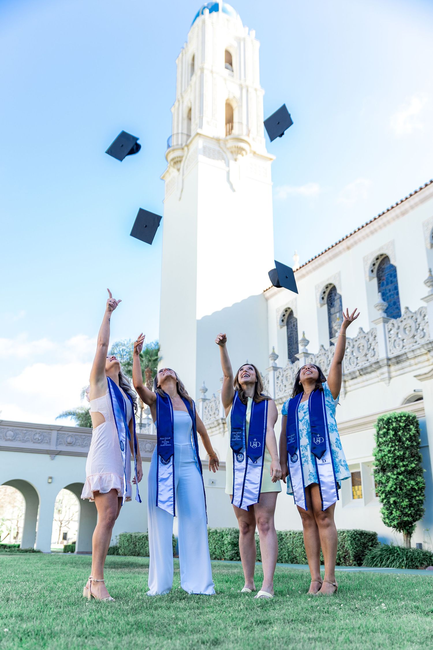 Graduates toss their mortarboards in front of a white church with blue domed tower during commencement celebration.